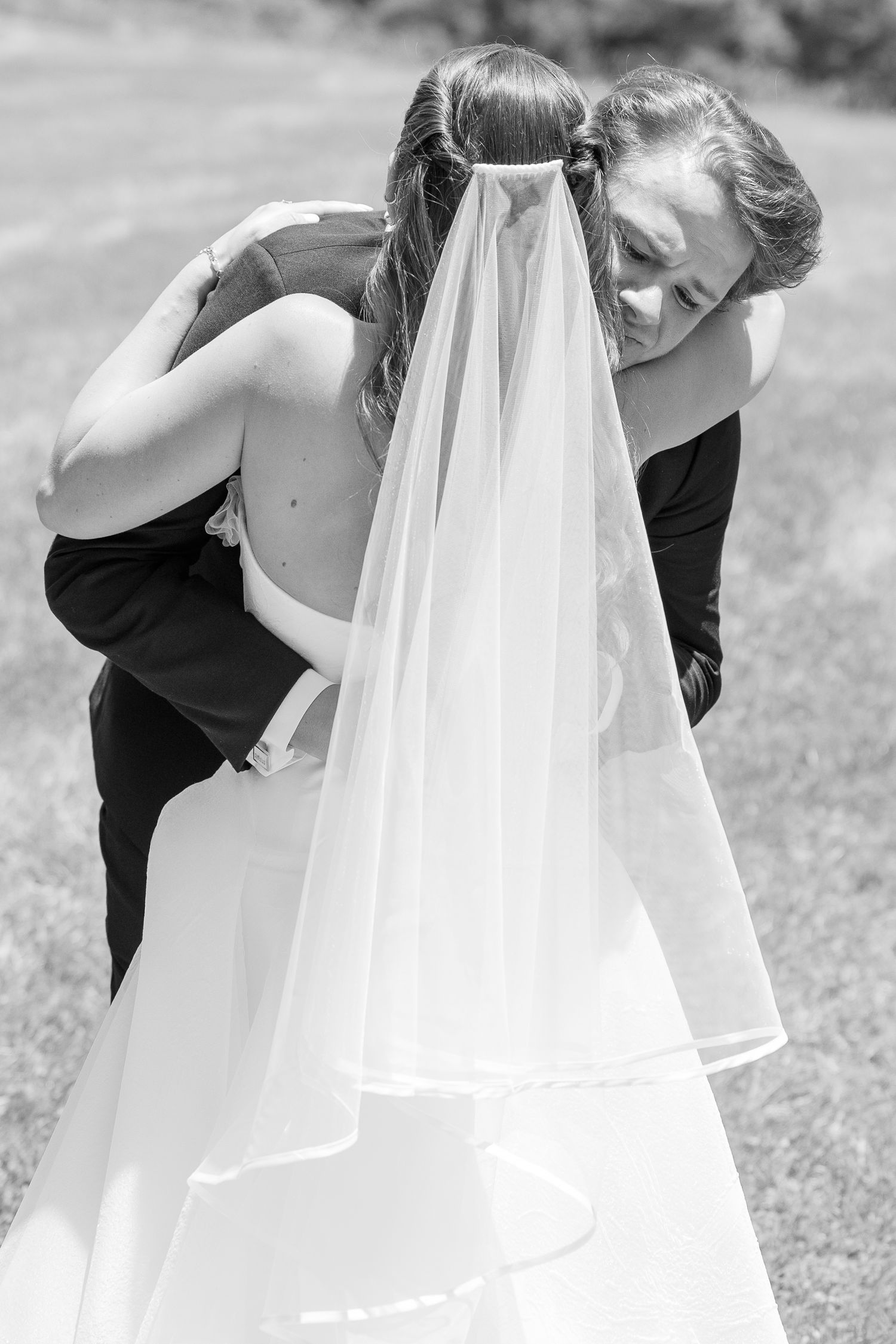 Black and white photo of a bride in a white gown and veil embracing a bridesmaid in a dark dress outdoors.
