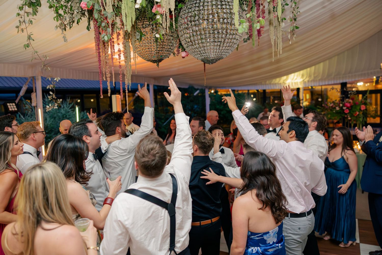 Wedding guests celebrate on the dance floor under crystal chandeliers in a white tent reception.
