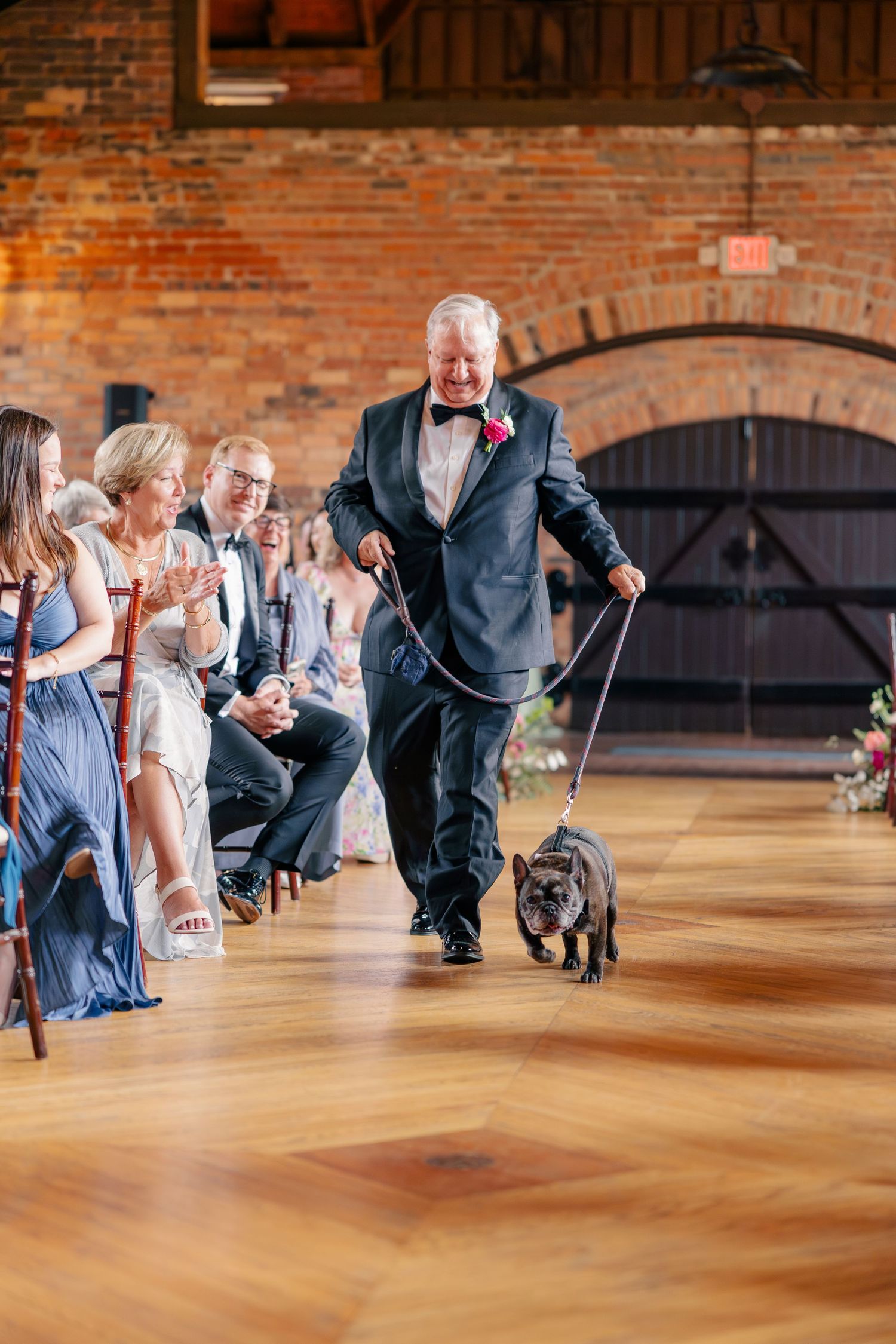 Elderly man in tuxedo walks French bulldog down wedding ceremony aisle past seated guests.