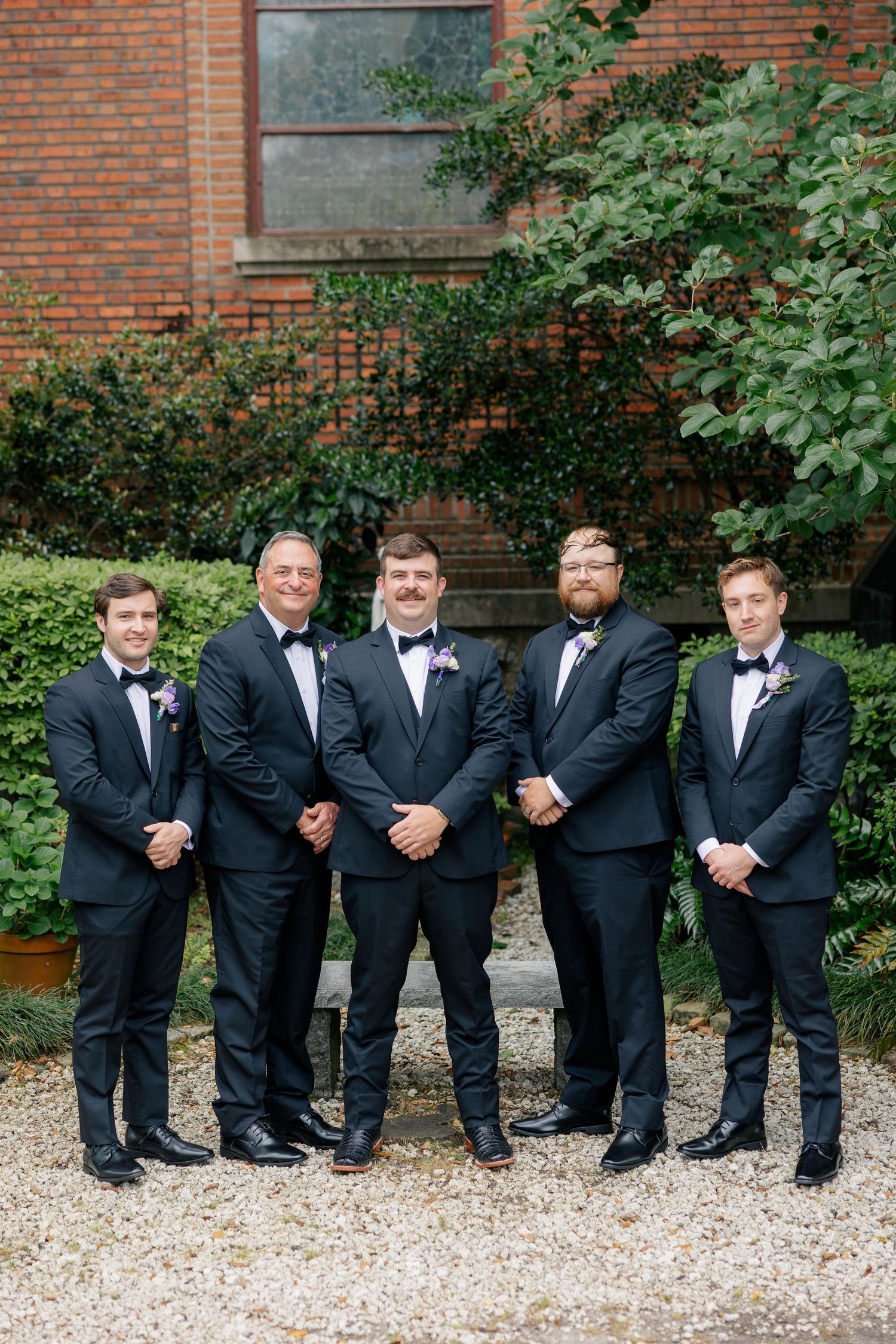 Five groomsmen in matching black tuxedos with bow ties pose outdoors in front of a brick building.