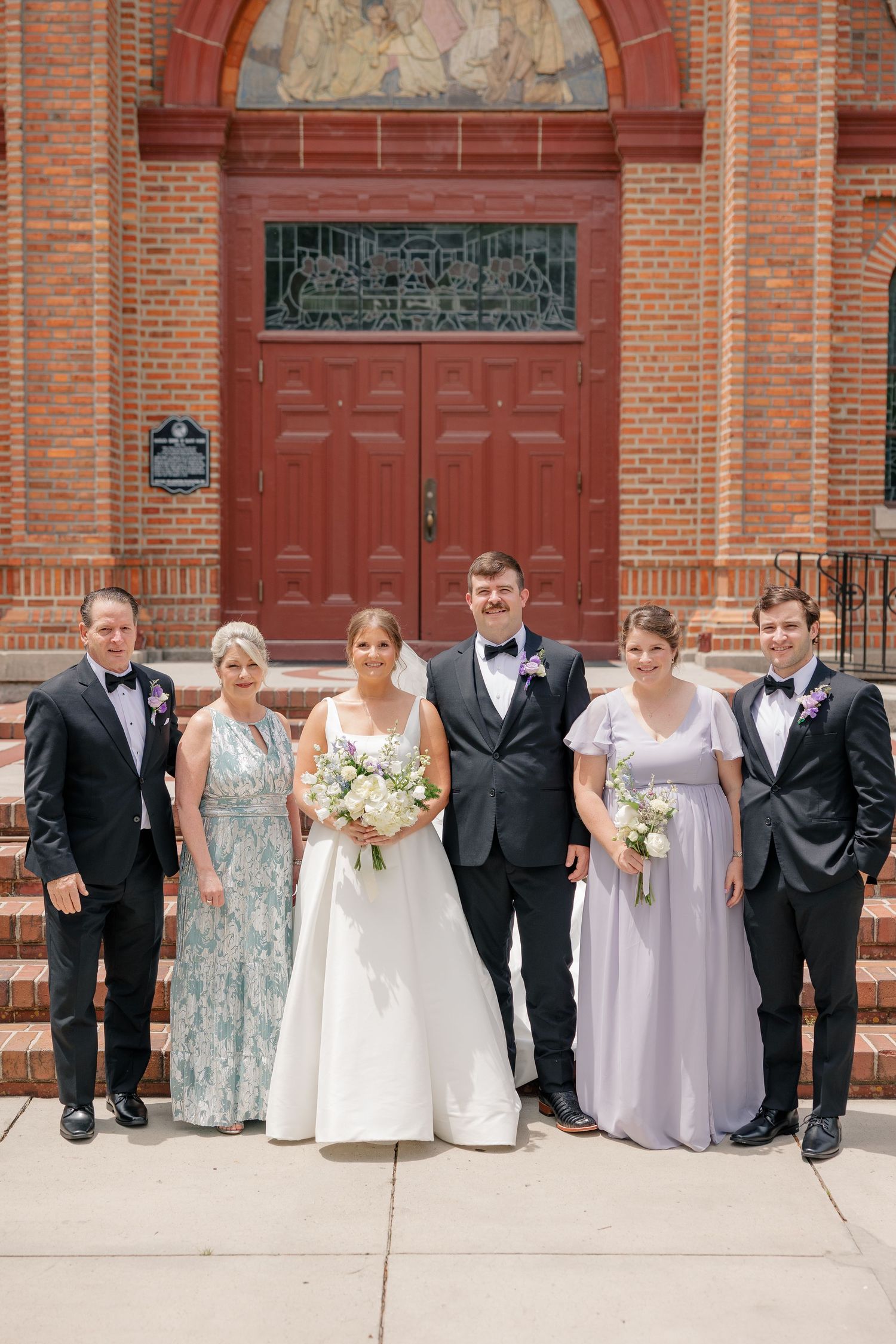 Wedding party of six posing in front of a red brick church with ornate wooden doors.