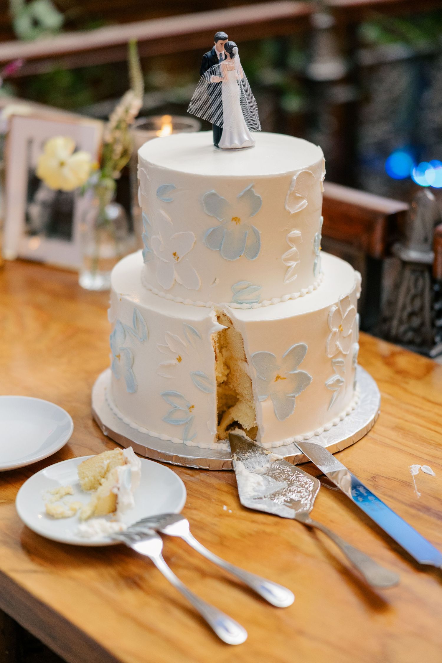 Two-tier white wedding cake with blue floral details and bride and groom topper, partially sliced on wooden table.