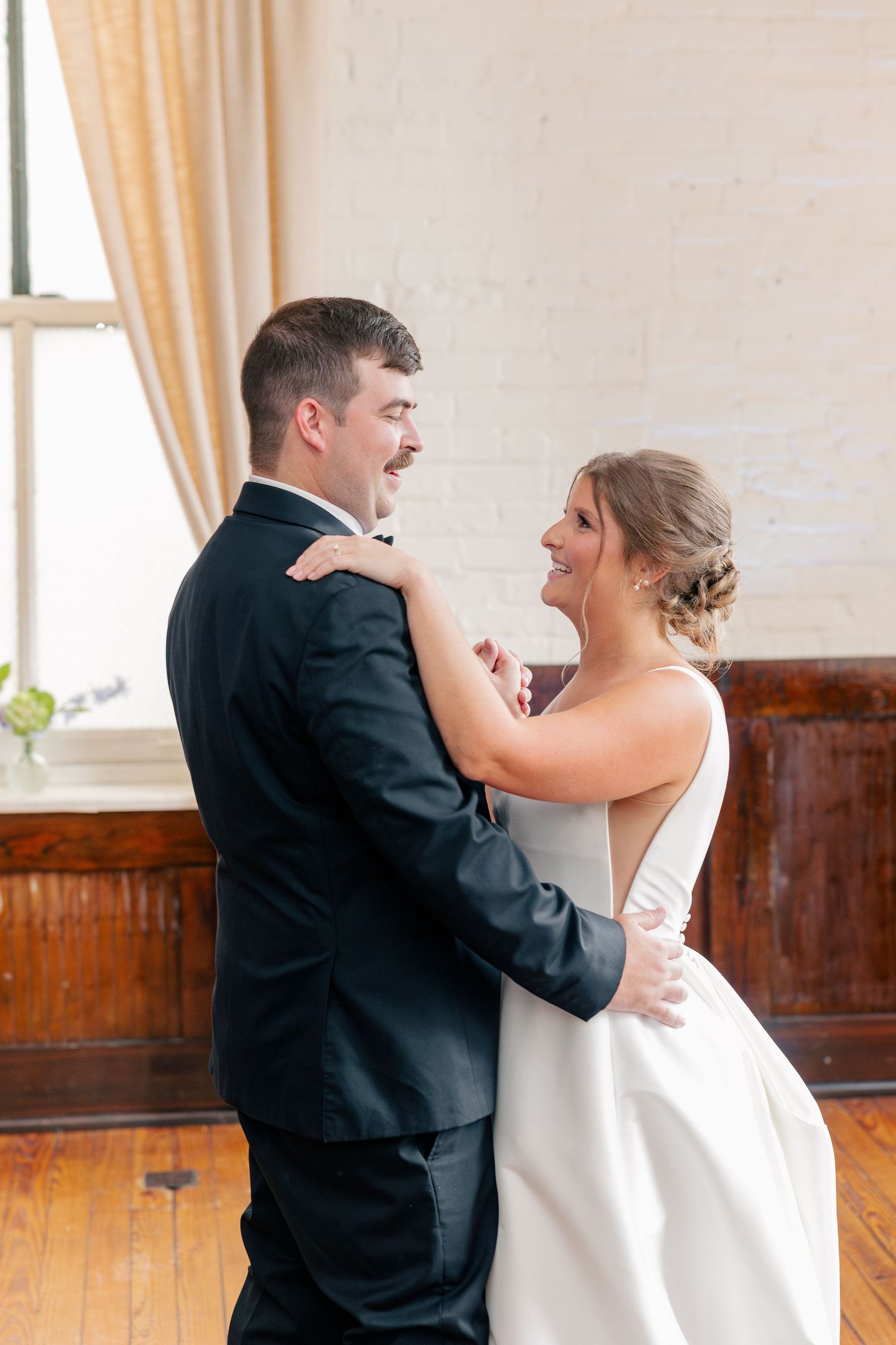 Bride and groom share joyful first dance embrace in rustic white brick venue with hardwood floors.