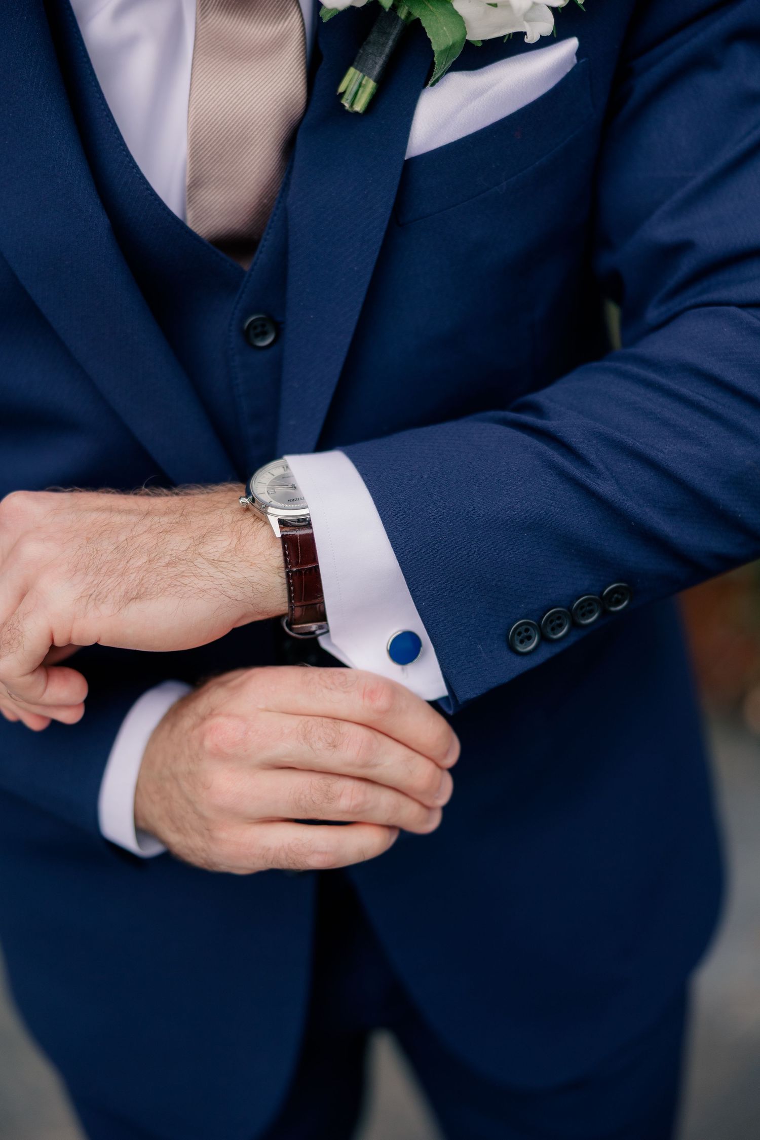 Groom in navy blue suit adjusting his watch and cufflinks, wearing a floral boutonniere on his lapel.