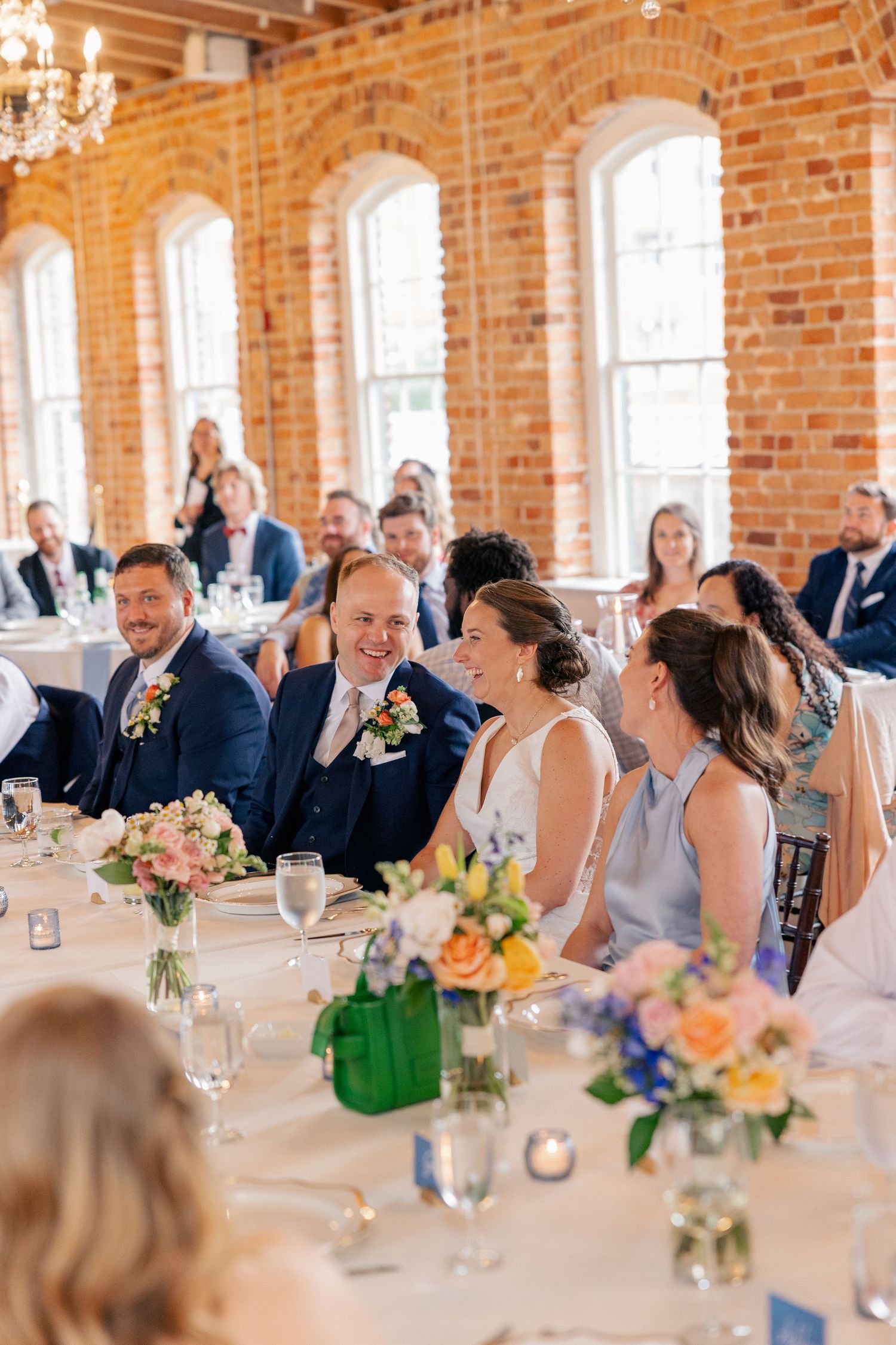 Bride and groom laugh with wedding party at head table in sunlit brick reception venue.