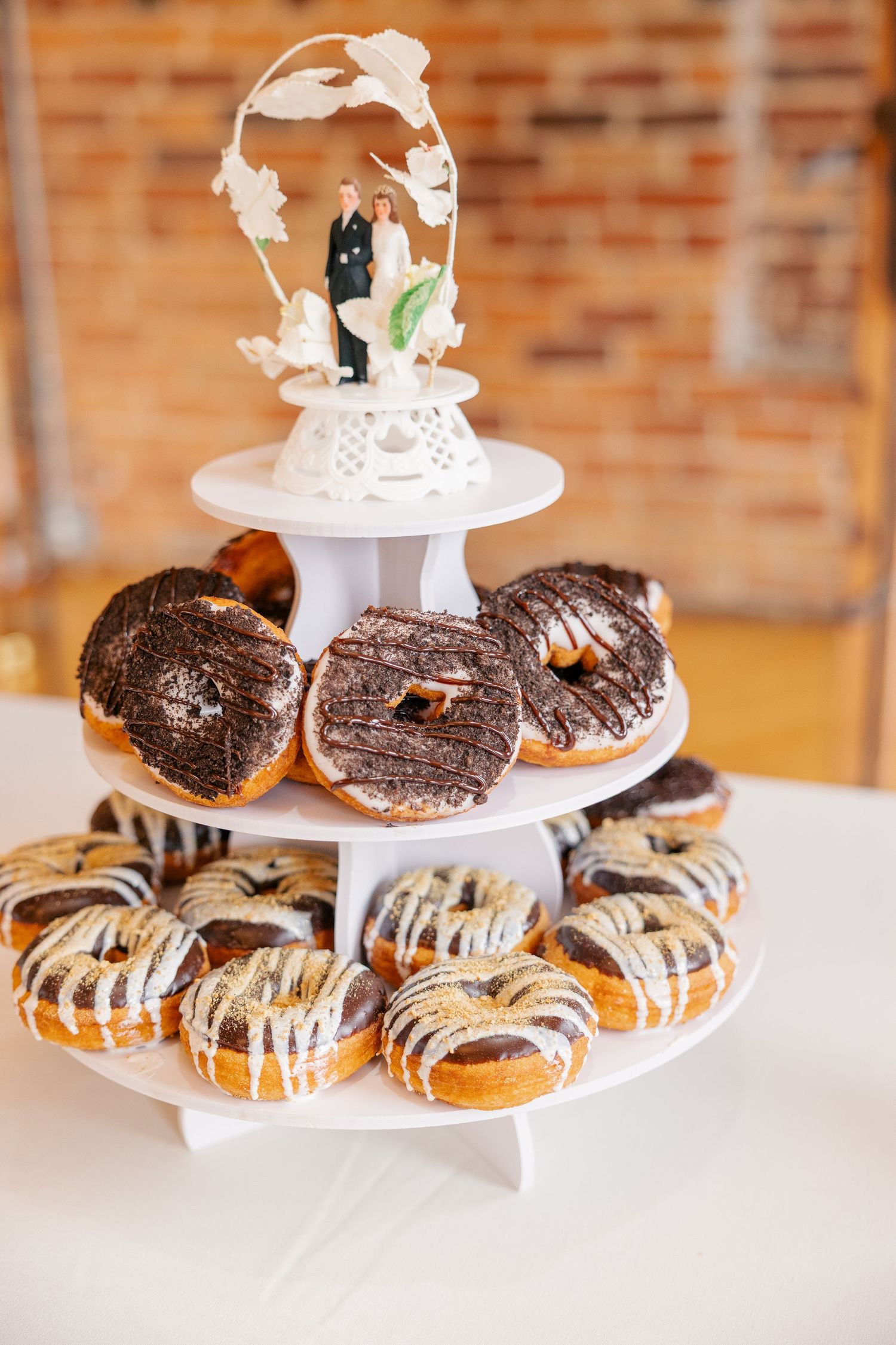 A tiered wedding donut display stand topped with bride and groom cake topper, featuring chocolate and glazed donuts.