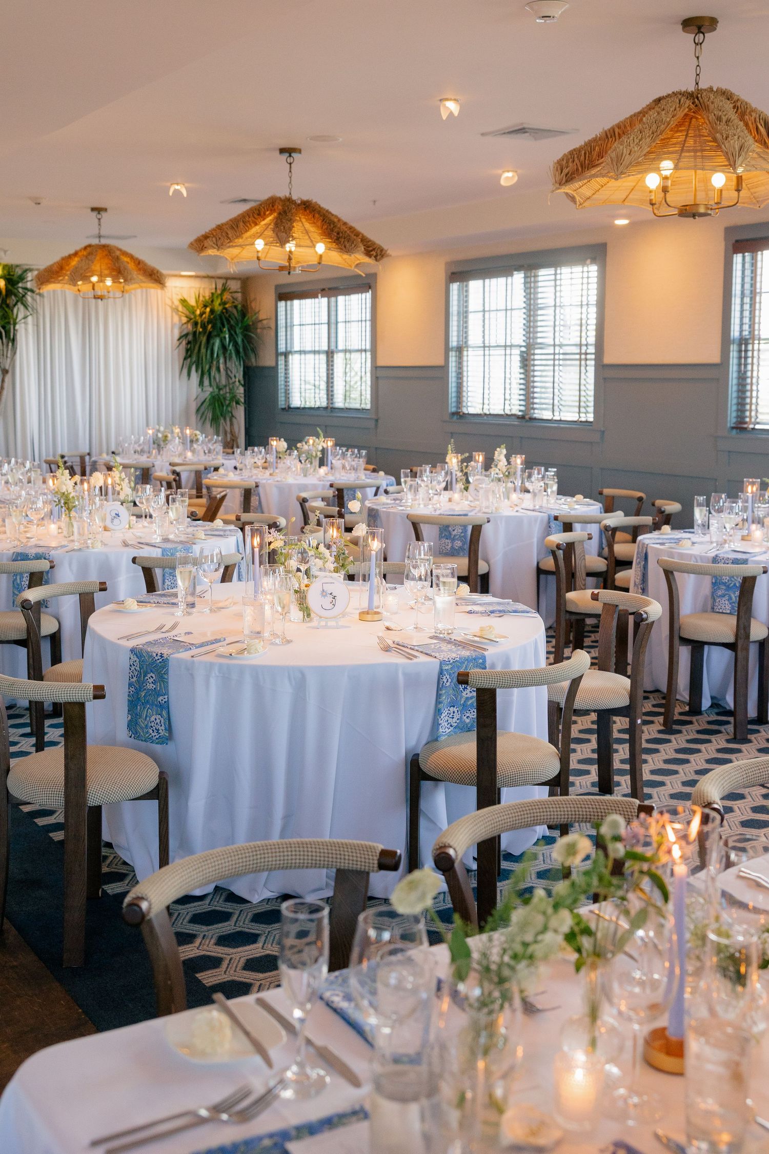 Romantic wedding reception venue with round tables, white linens, blue accents, candlelight, and woven pendant lights.