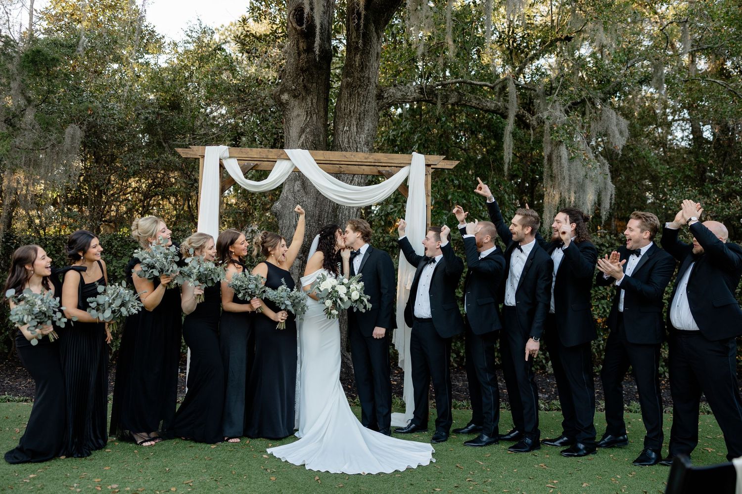Wedding party poses for fun group photo under draped outdoor ceremony arch.
