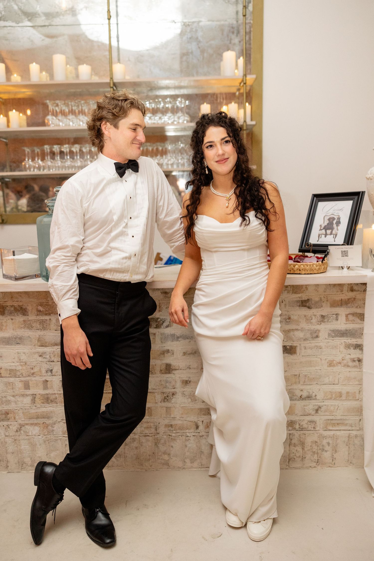 Bride and groom pose casually at indoor bar area with exposed brick and glassware display.