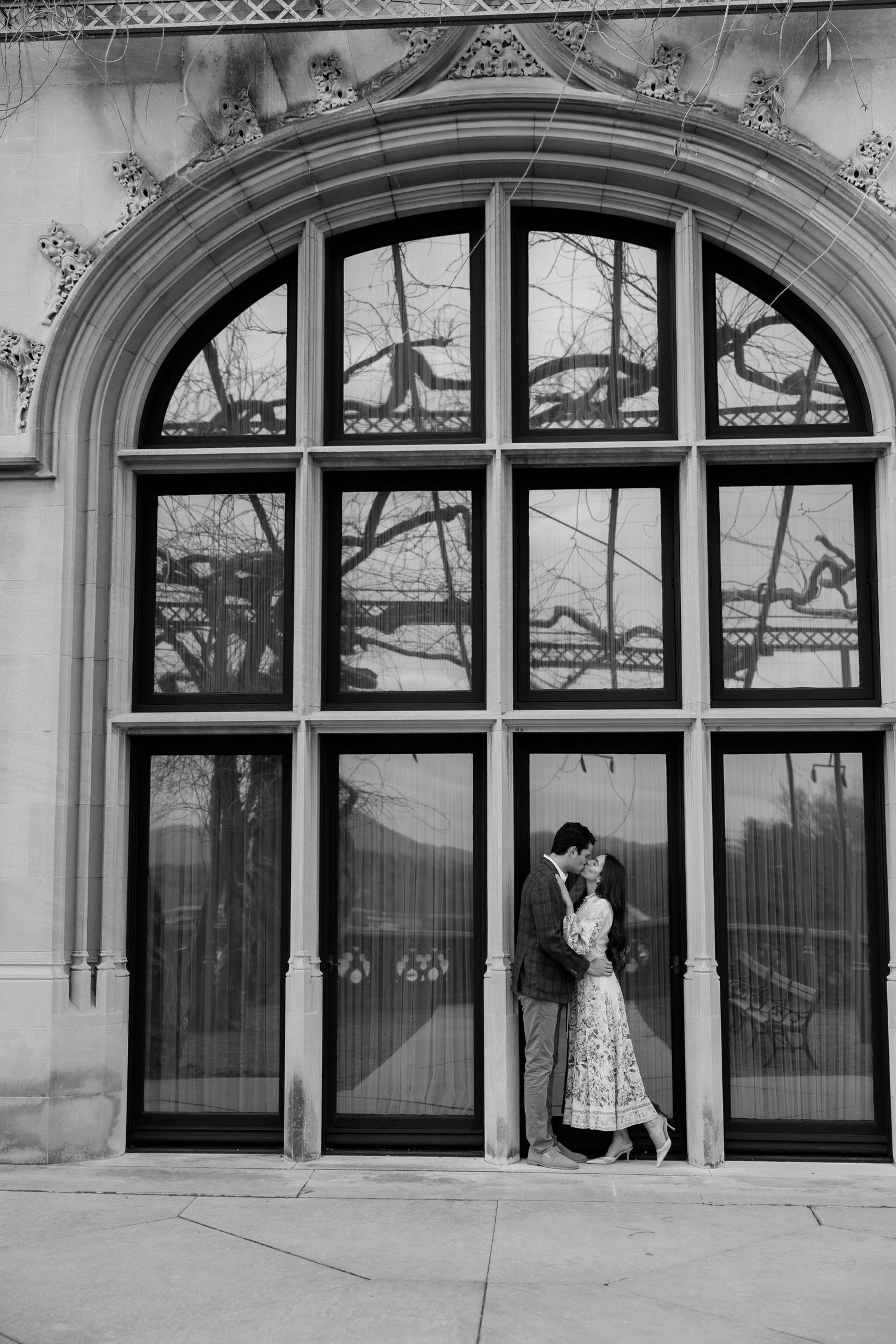 A couple stands intimately by an ornate arched window with gothic architectural details in black and white.