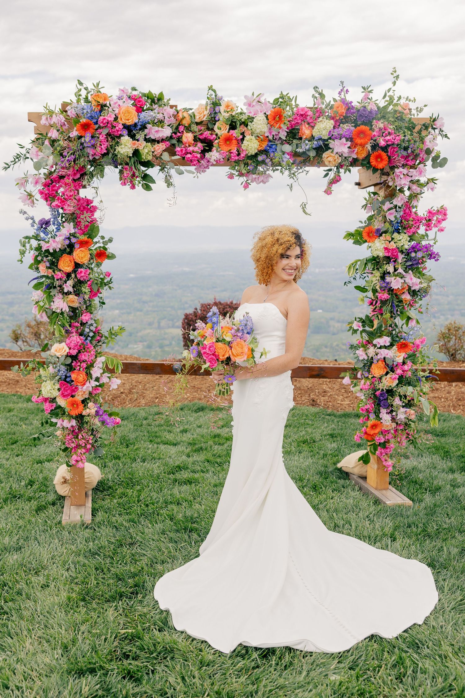 Smiling bride in white gown holds bouquet under a stunning colorful floral arch with mountain views behind her.