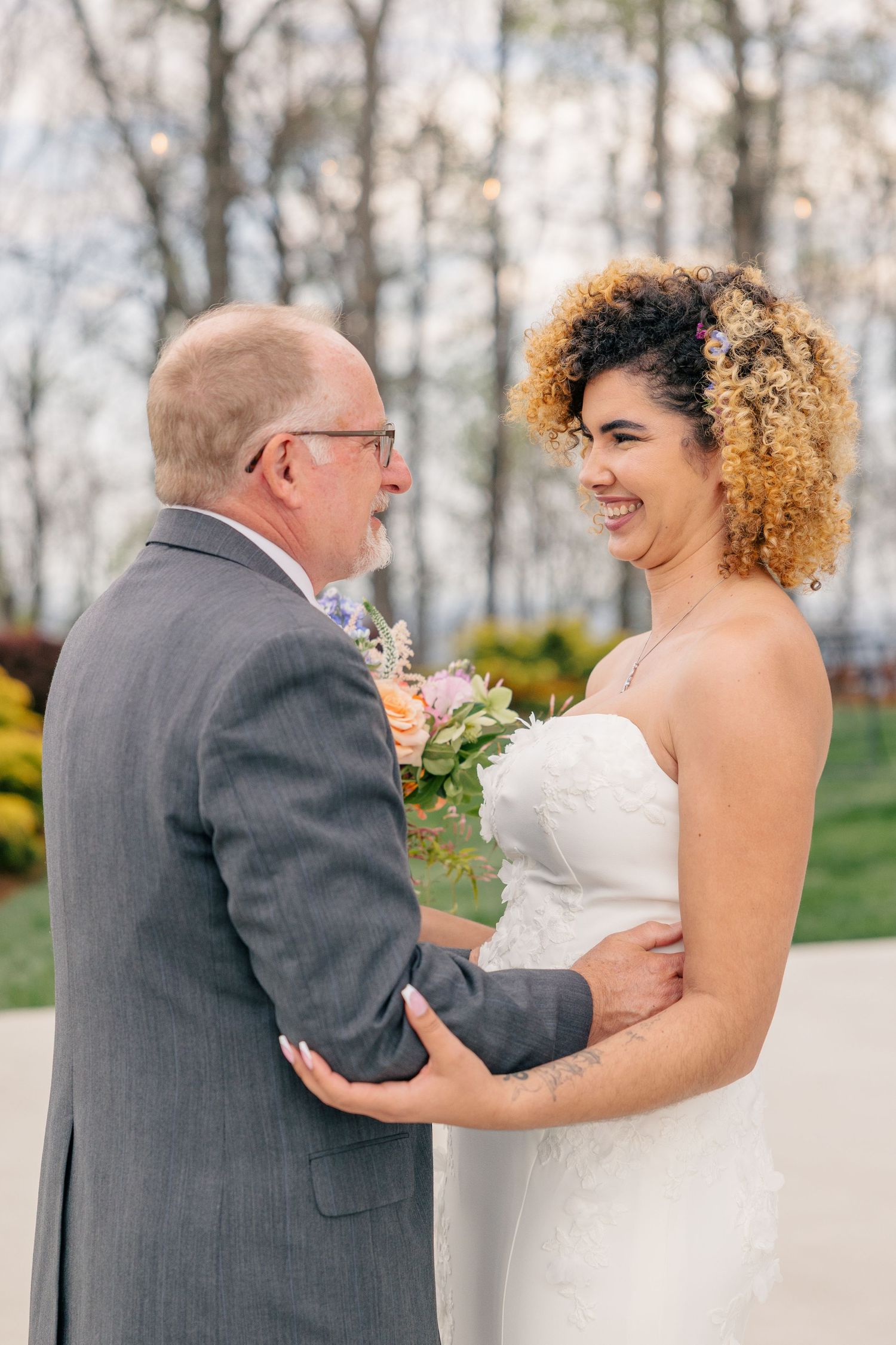 Bride in white strapless gown smiles at older man in gray suit during wedding first look.