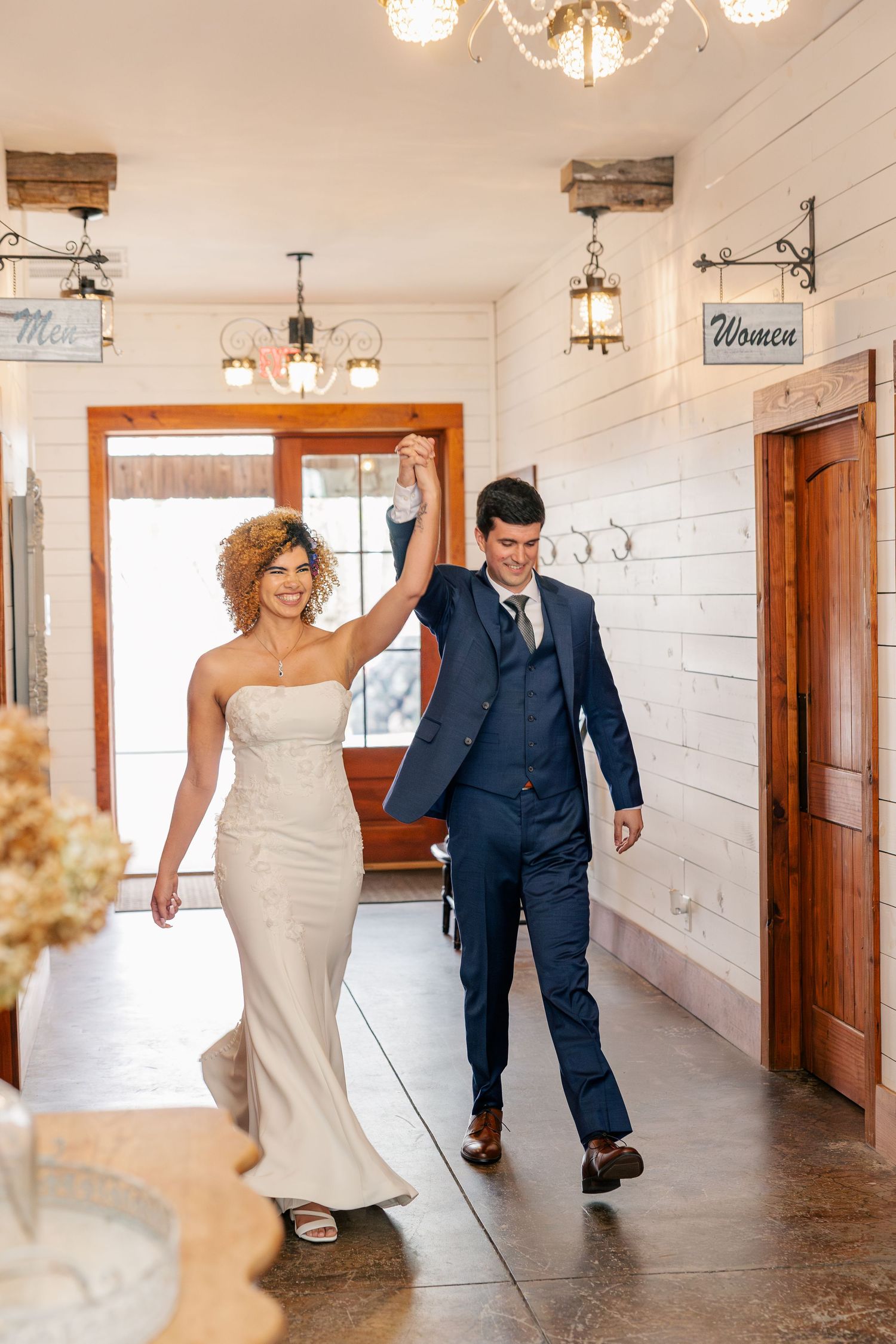 Joyful newlyweds making a grand entrance hand in hand into a rustic wedding venue with warm lighting.