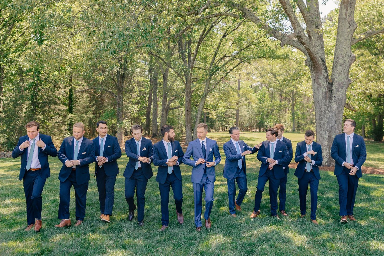 Groomsmen in navy suits adjust ties while walking across green lawn with trees in background.