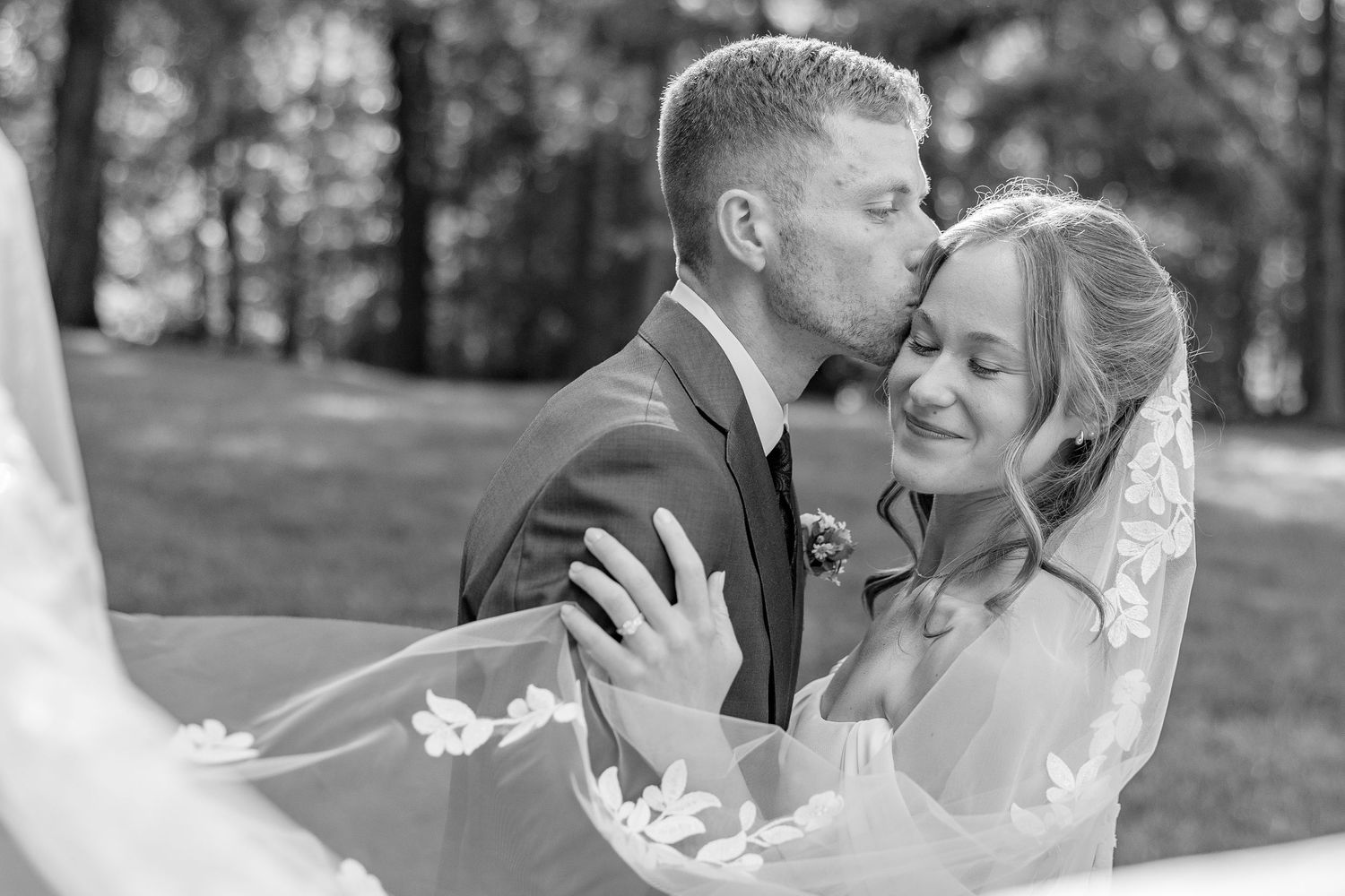 Black and white photo of groom kissing bride on cheek as veil flows in wind.