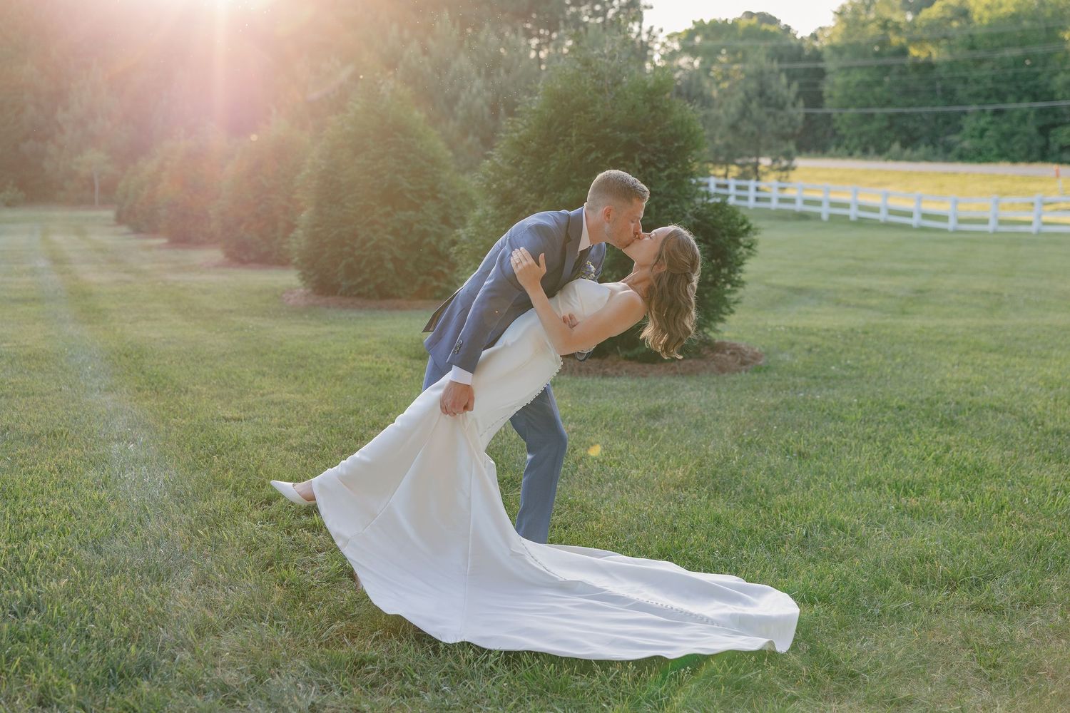 Bride and groom sharing a romantic dip kiss on lush green lawn during golden hour sunset at outdoor wedding.