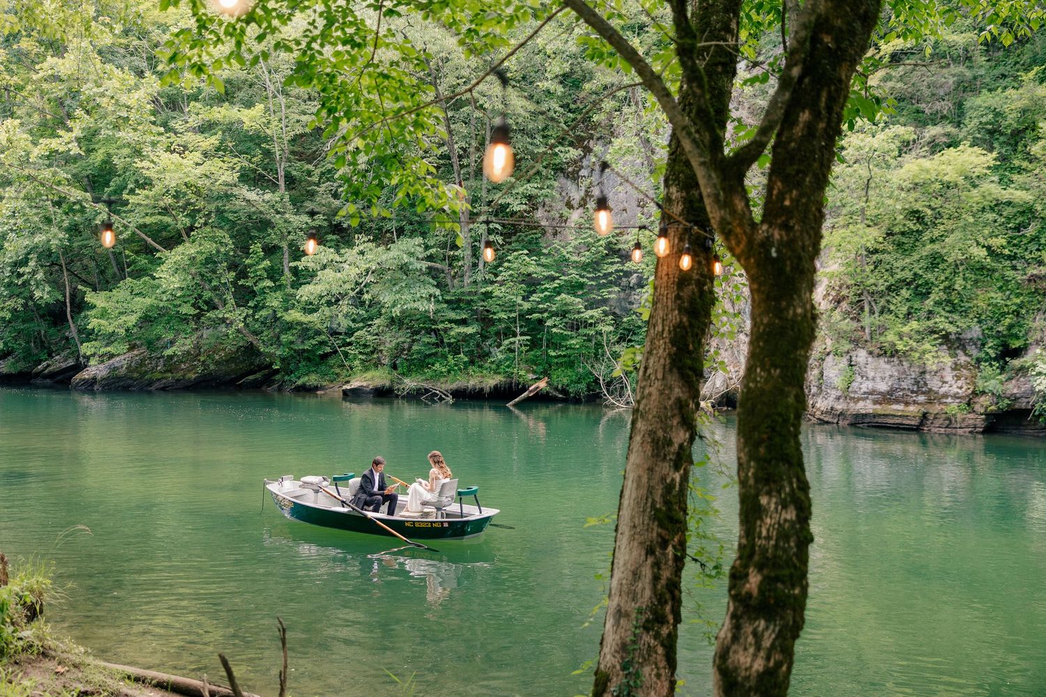 Couple sitting in a small boat on a serene green river surrounded by lush forest trees with hanging Edison lights.