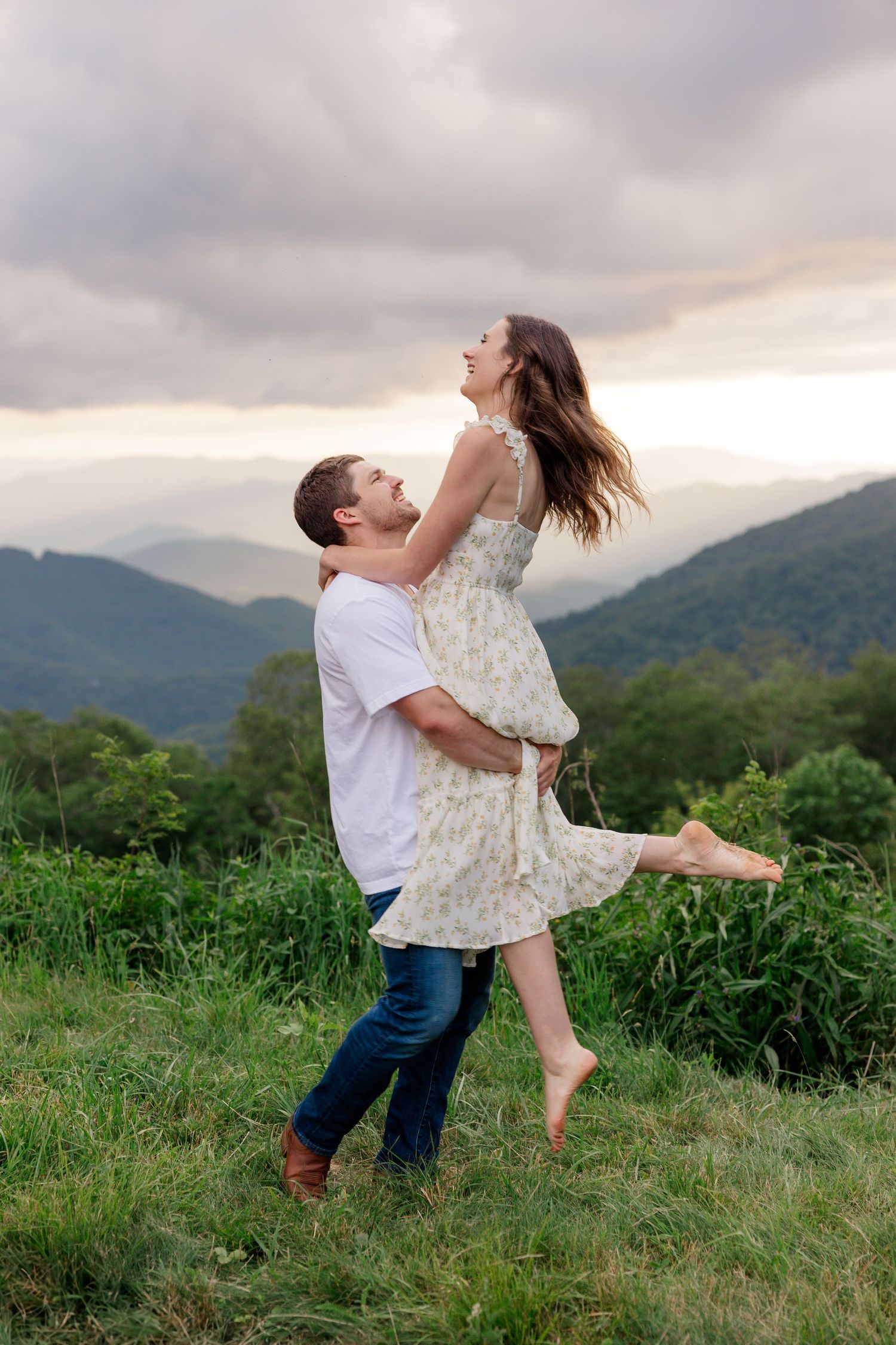 A playful couple spins and dances together in a grassy mountain field at sunset.