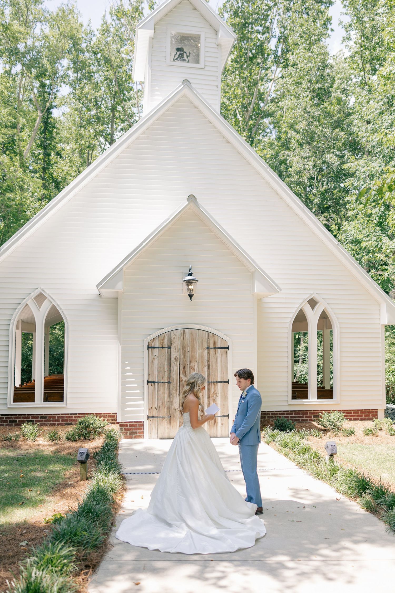 Bride and groom share vows outside a charming white chapel surrounded by trees.