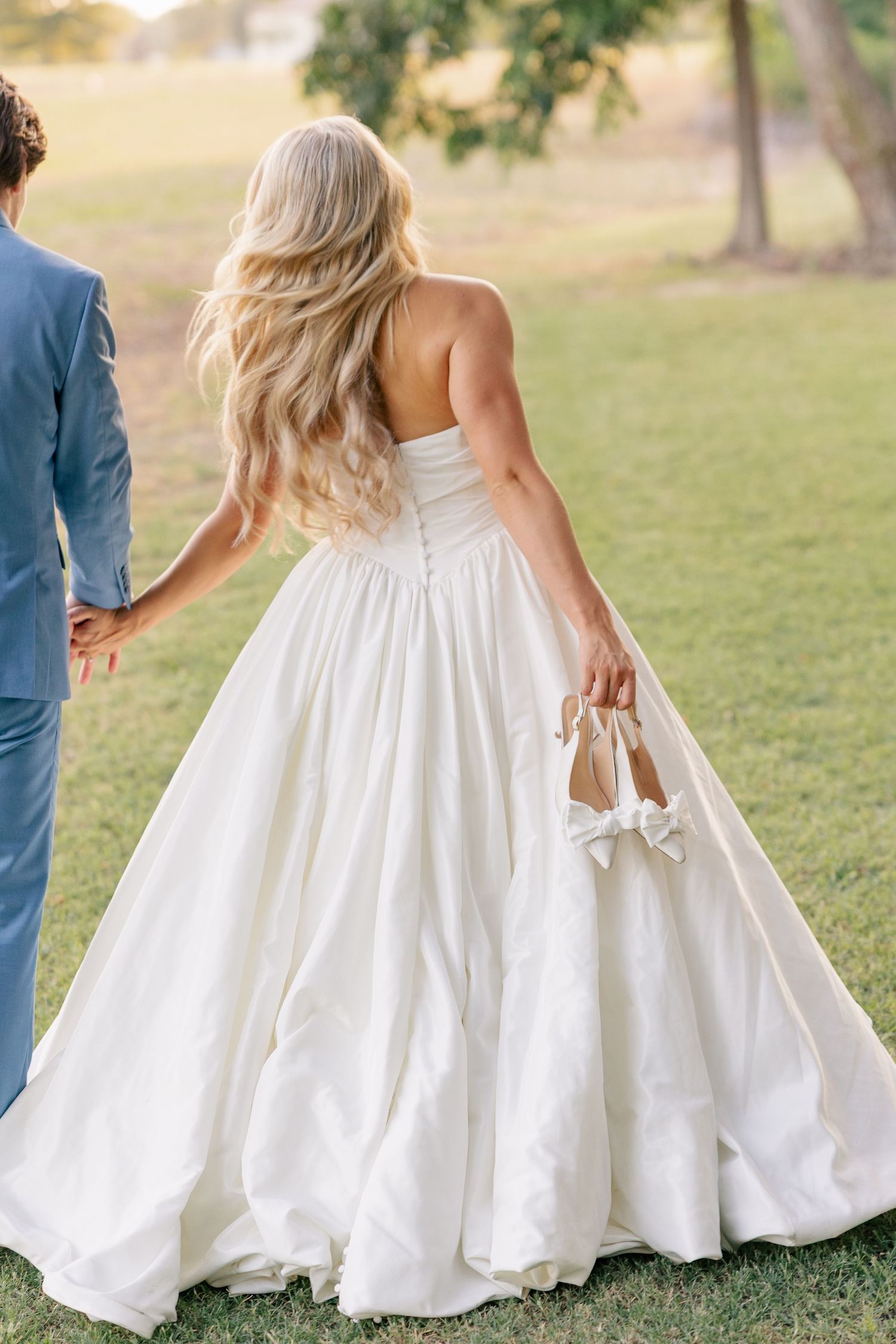 Bride carries white heels while holding grooms hand walking through green lawn in ballgown.