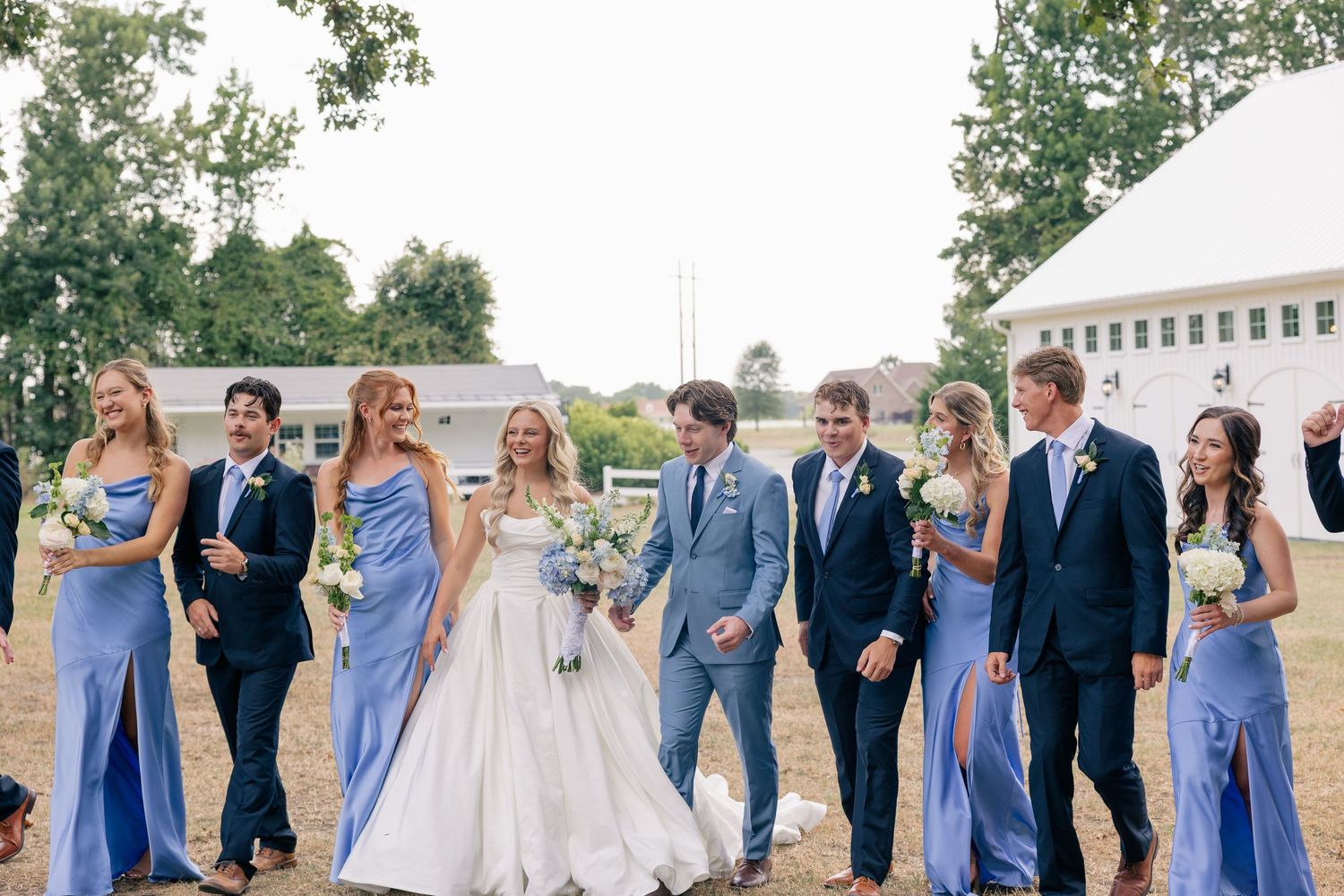 Wedding party walks outdoors, bride in white gown with bridesmaids in blue dresses and groomsmen in navy suits.