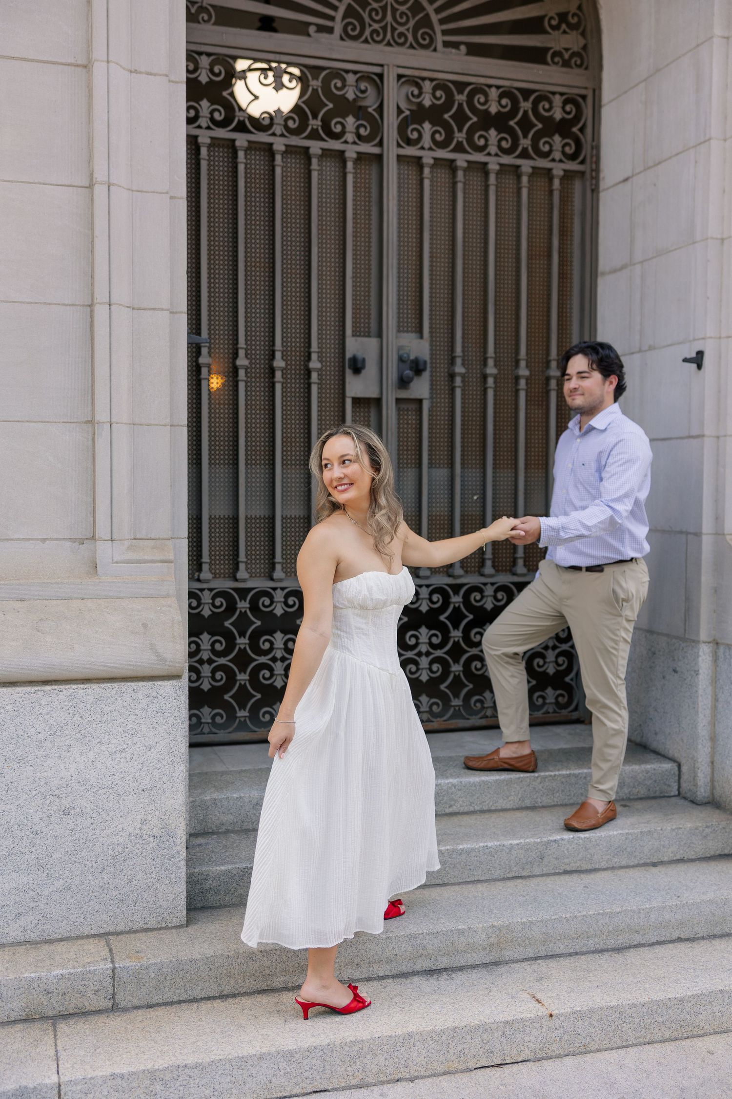 Smiling bride in white strapless dress and red heels leads groom by hand down ornate building steps.