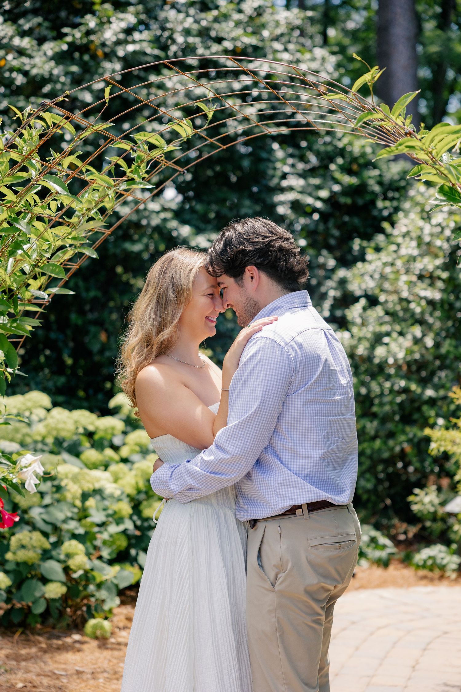 Couple embracing tenderly under a lush green garden arch, sharing a romantic moment outdoors on a sunny day.