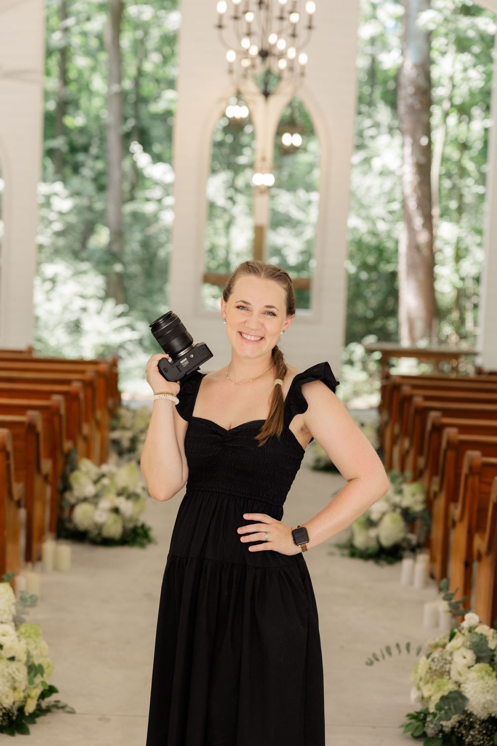 Wedding photographer in black dress posing with camera in elegant chapel setting.