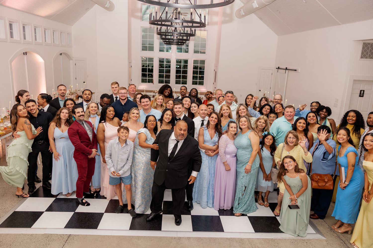 Large wedding party group photo on black and white checkered floor in elegant venue.