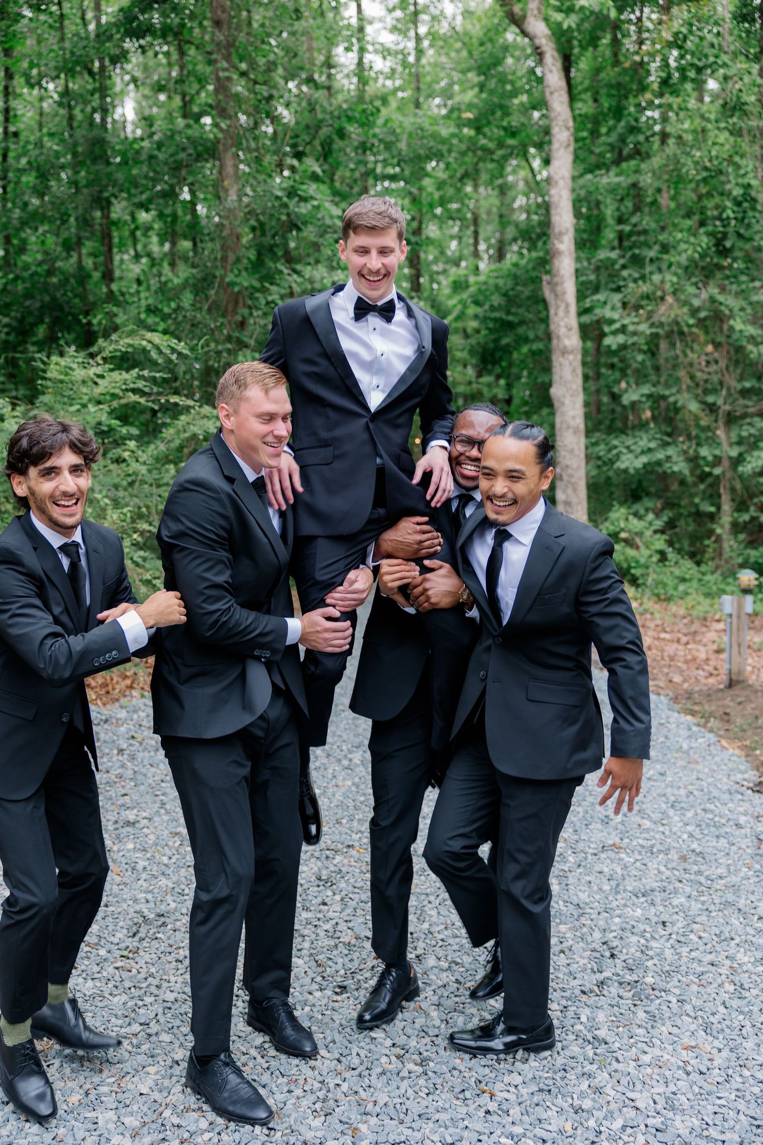 Wedding party in black tuxedos posing playfully on a gravel path surrounded by trees.