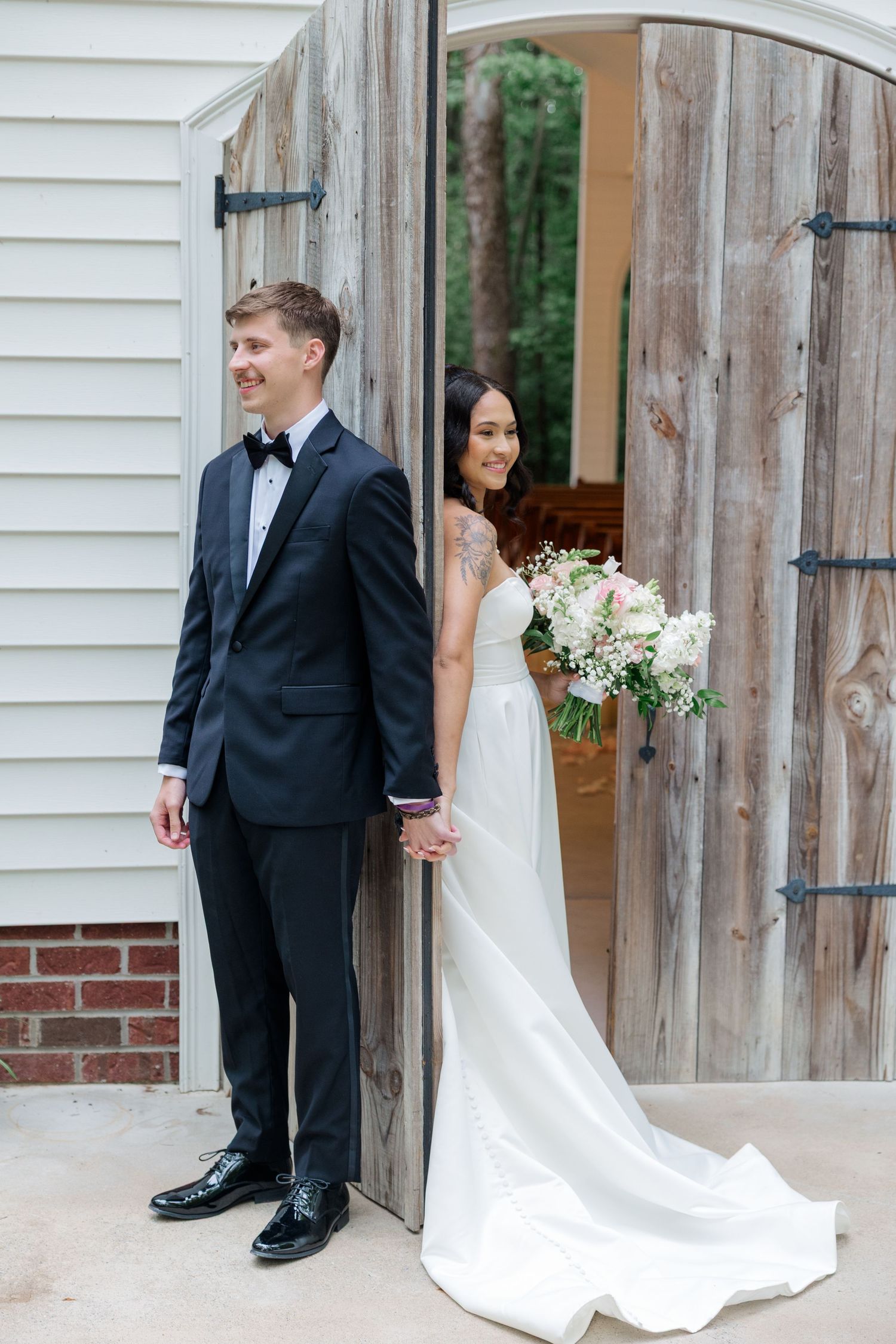 Wedding couple shares a moment by rustic wooden doors of white chapel.