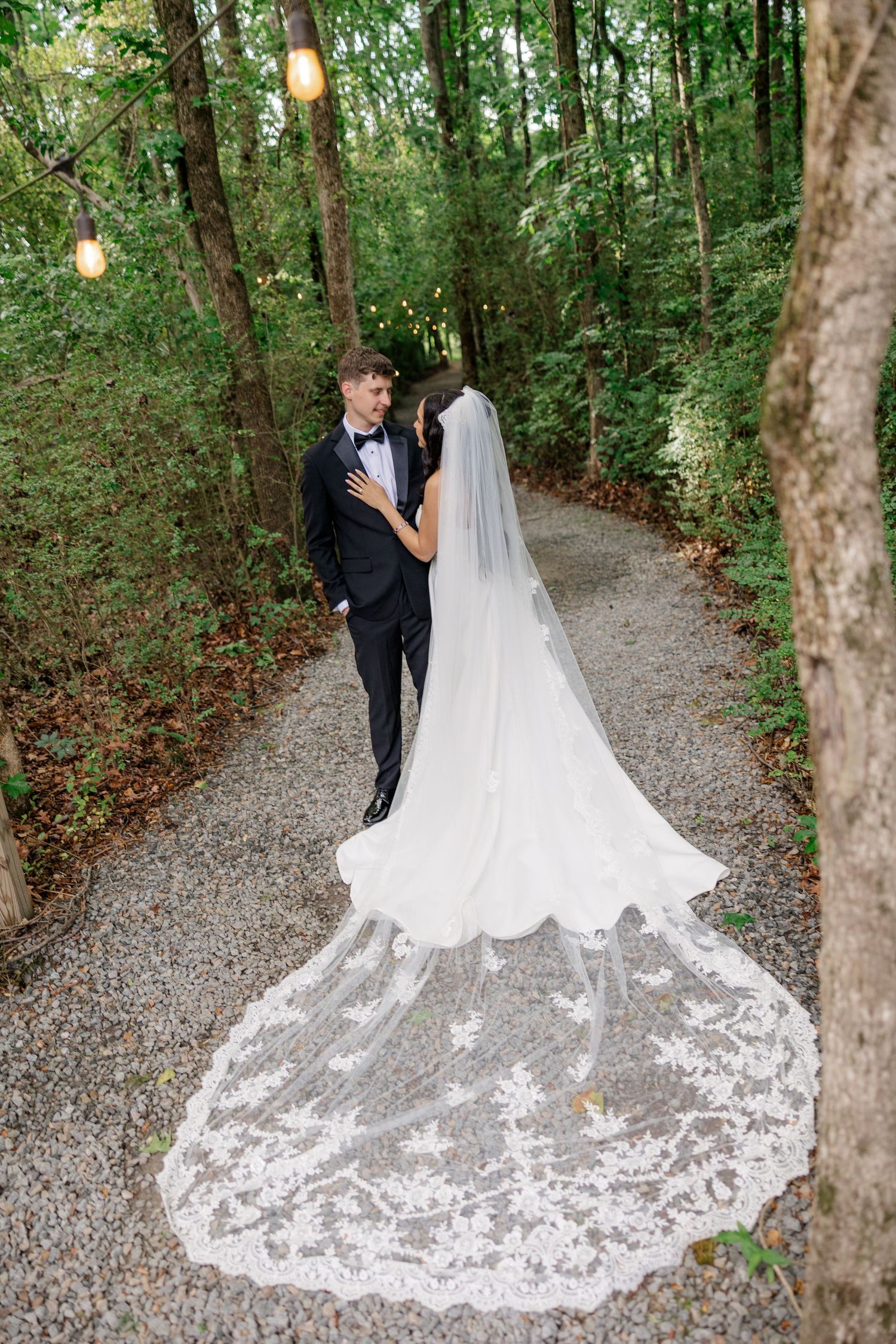 A romantic wedding photo shoot on a wooded path with string lights overhead and a flowing lace wedding gown train.