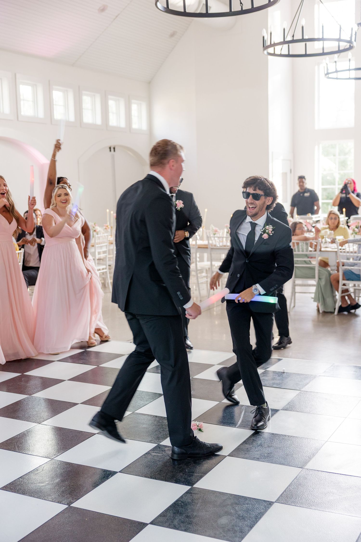 Wedding party dances on black and white checkered floor in bright reception hall.