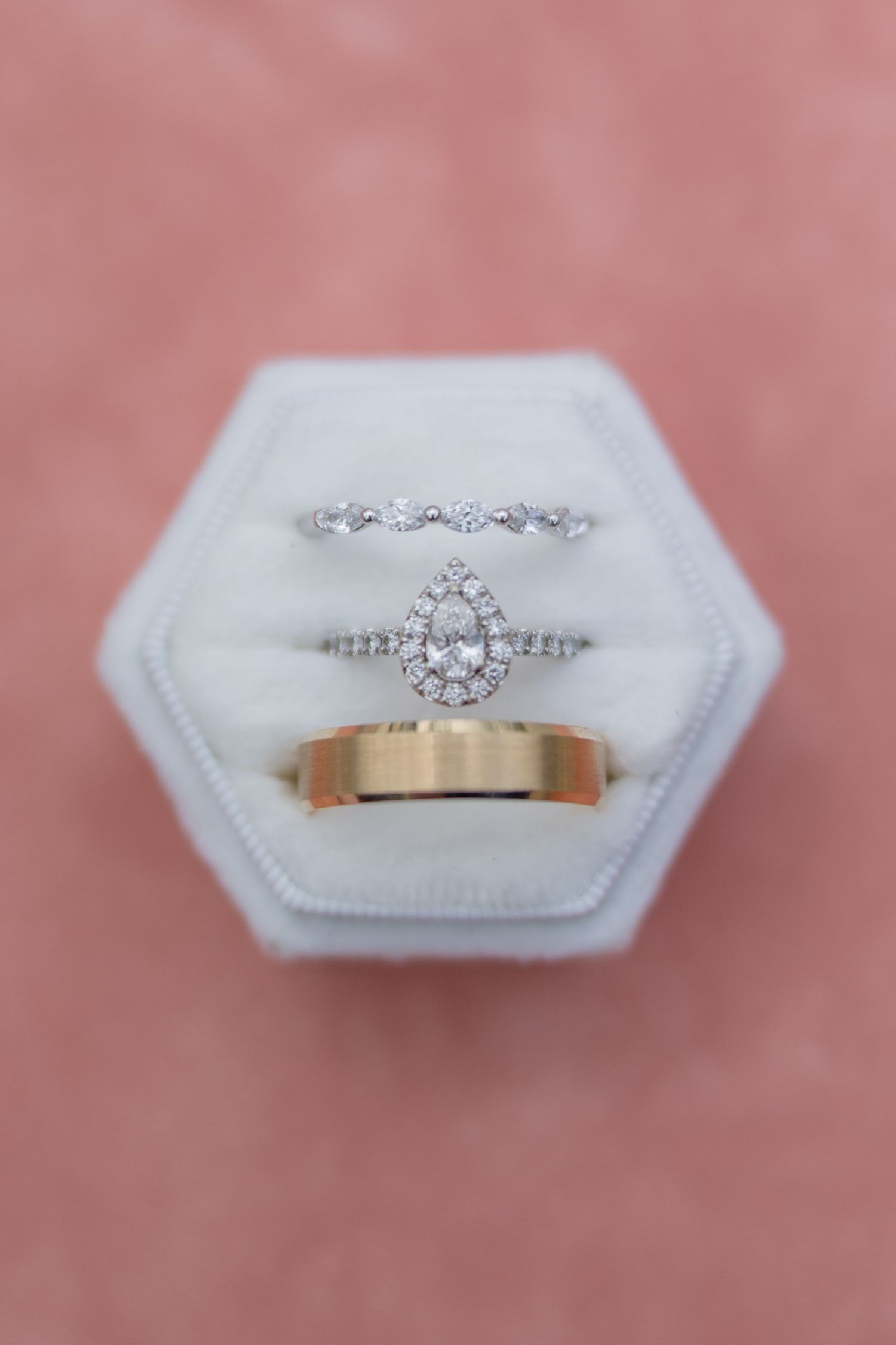 A collection of wedding rings and engagement rings displayed on white hexagonal cushions against a pink background.