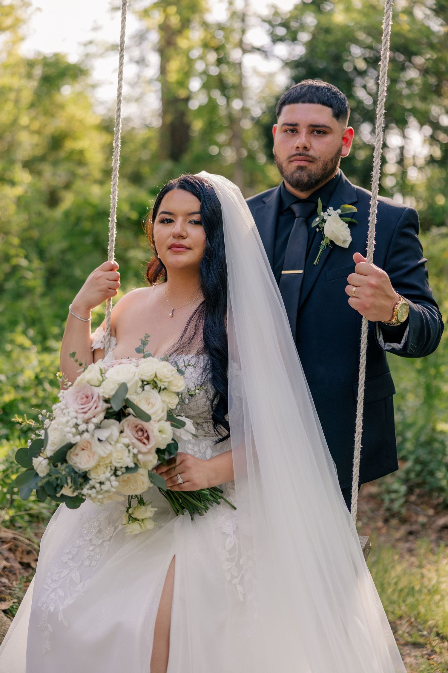 Couple on a swing in a forest setting with bride holding a white and pink bouquet.