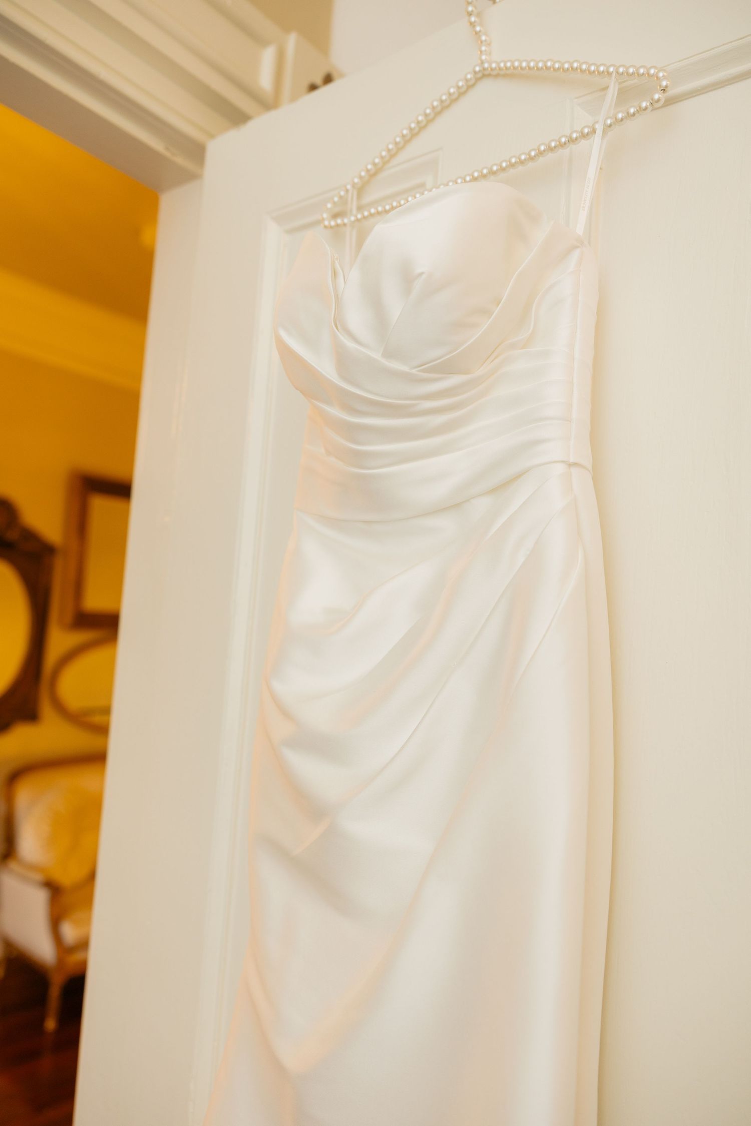 White satin wedding dress hanging against cream colored wall showing elegant draping details.