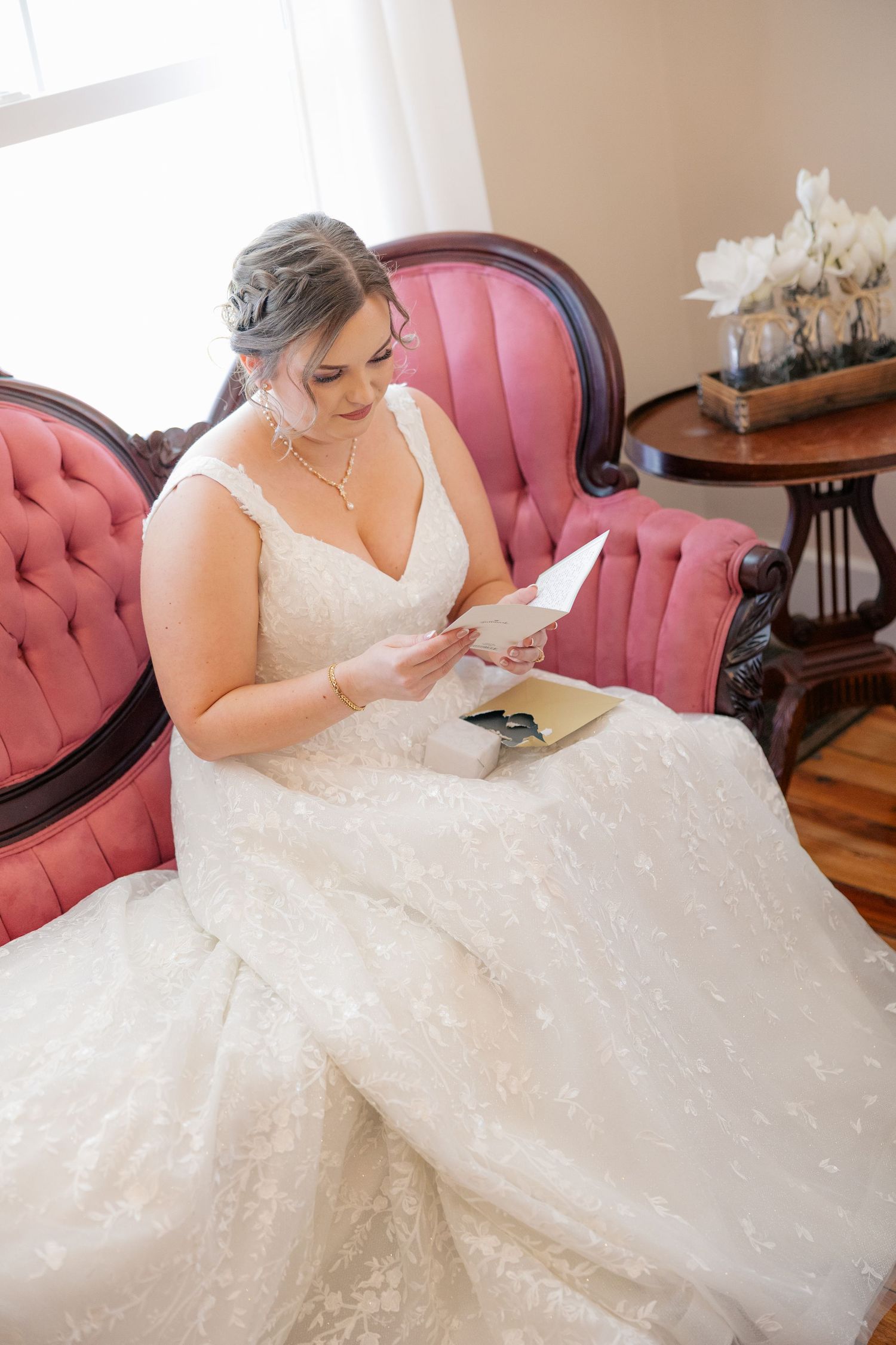A bride in a stunning lace gown sits on a pink vintage couch reading.