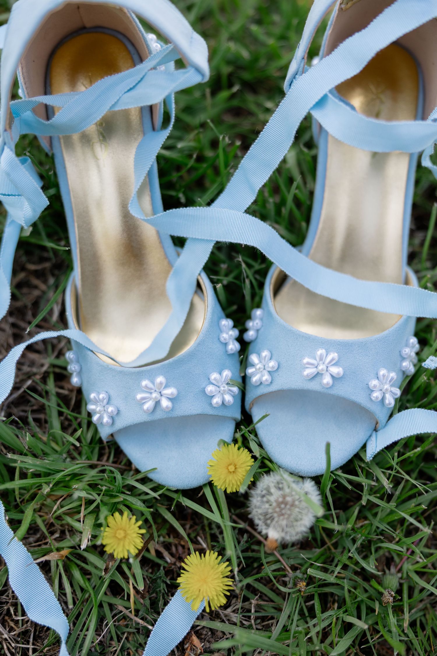Light blue bridal shoes with floral details resting on grass with dandelions.