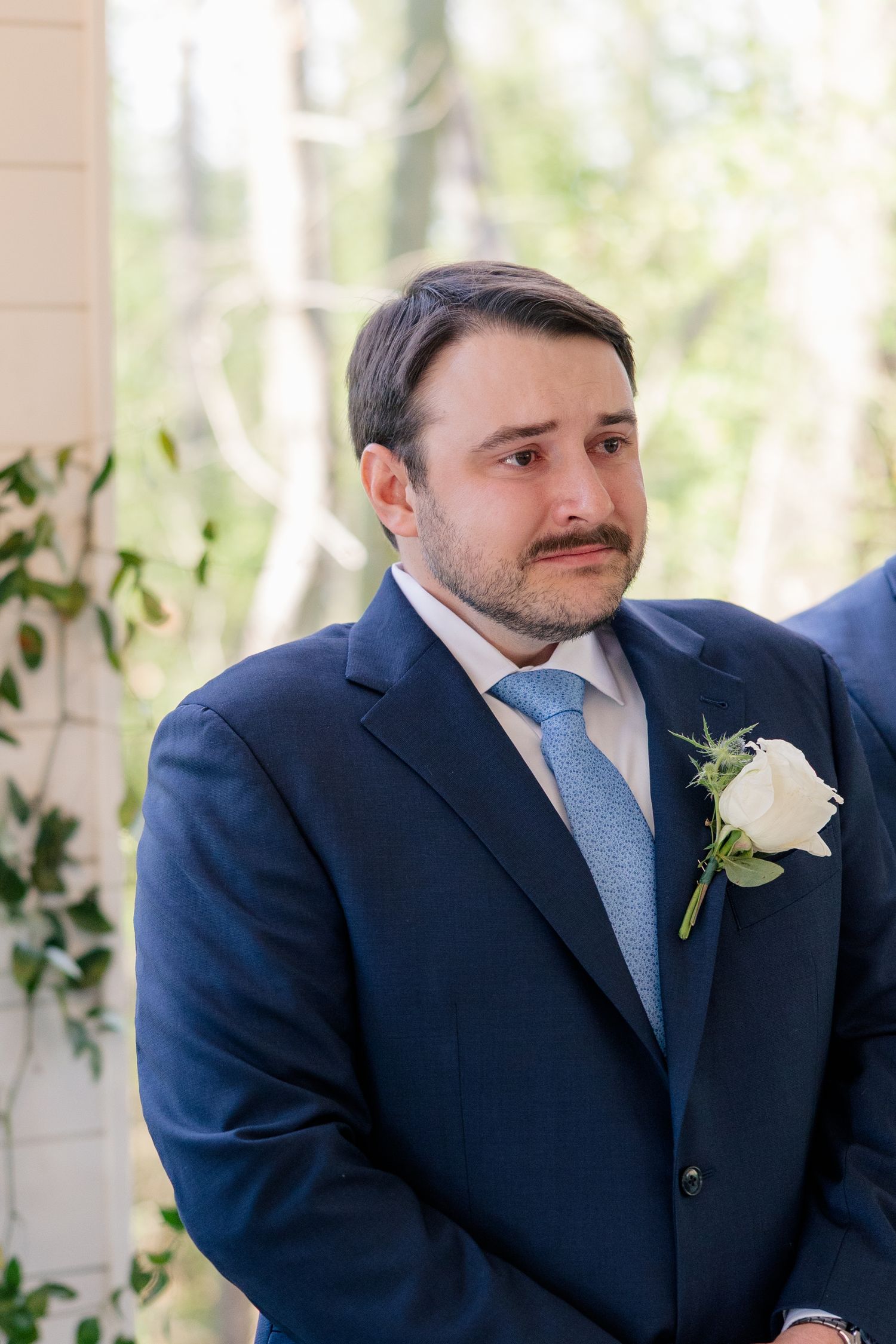 Groom wearing a navy suit with light blue tie and white boutonniere in an outdoor setting.