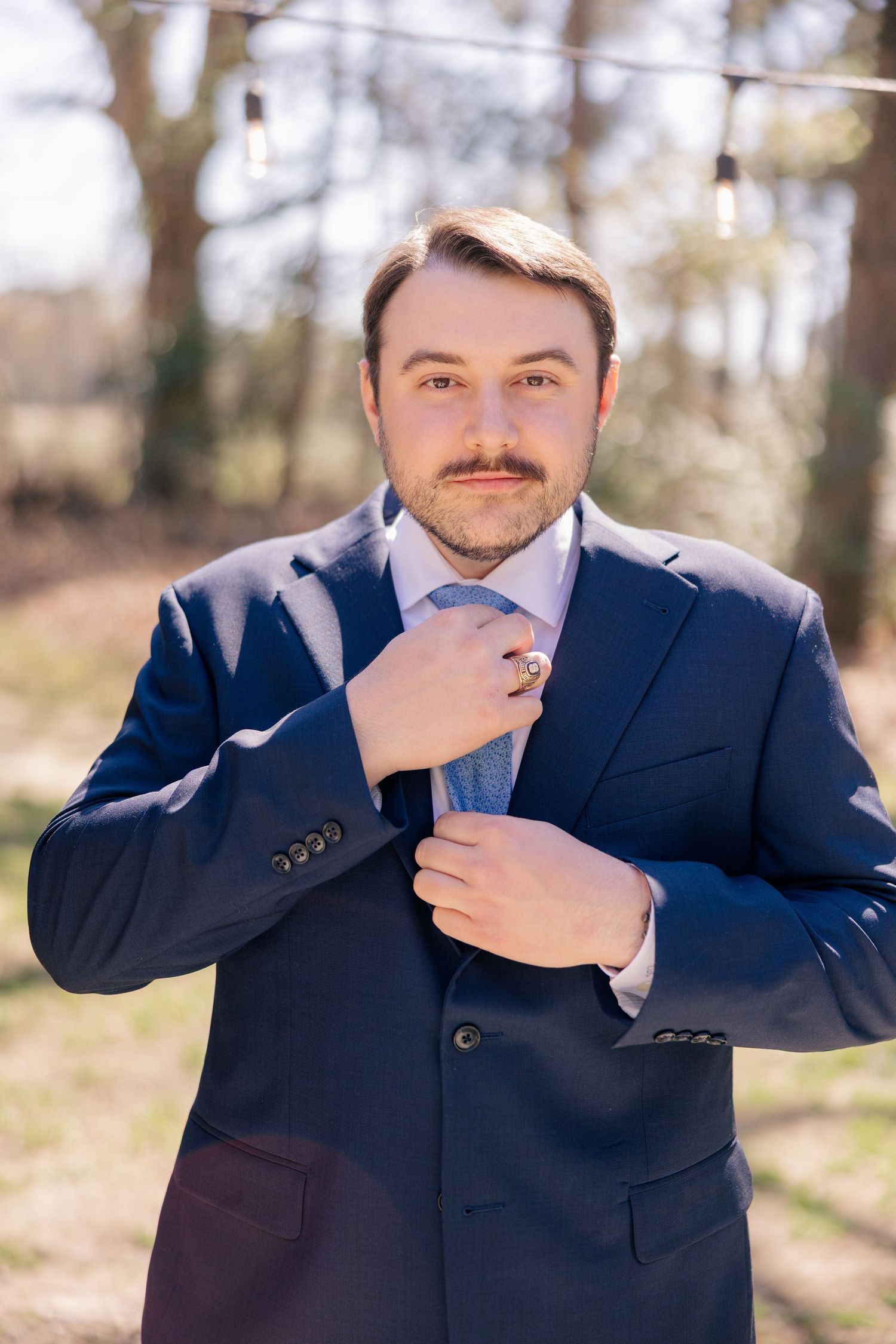Groom adjusts his navy blue suit and tie while standing outdoors in natural lighting.