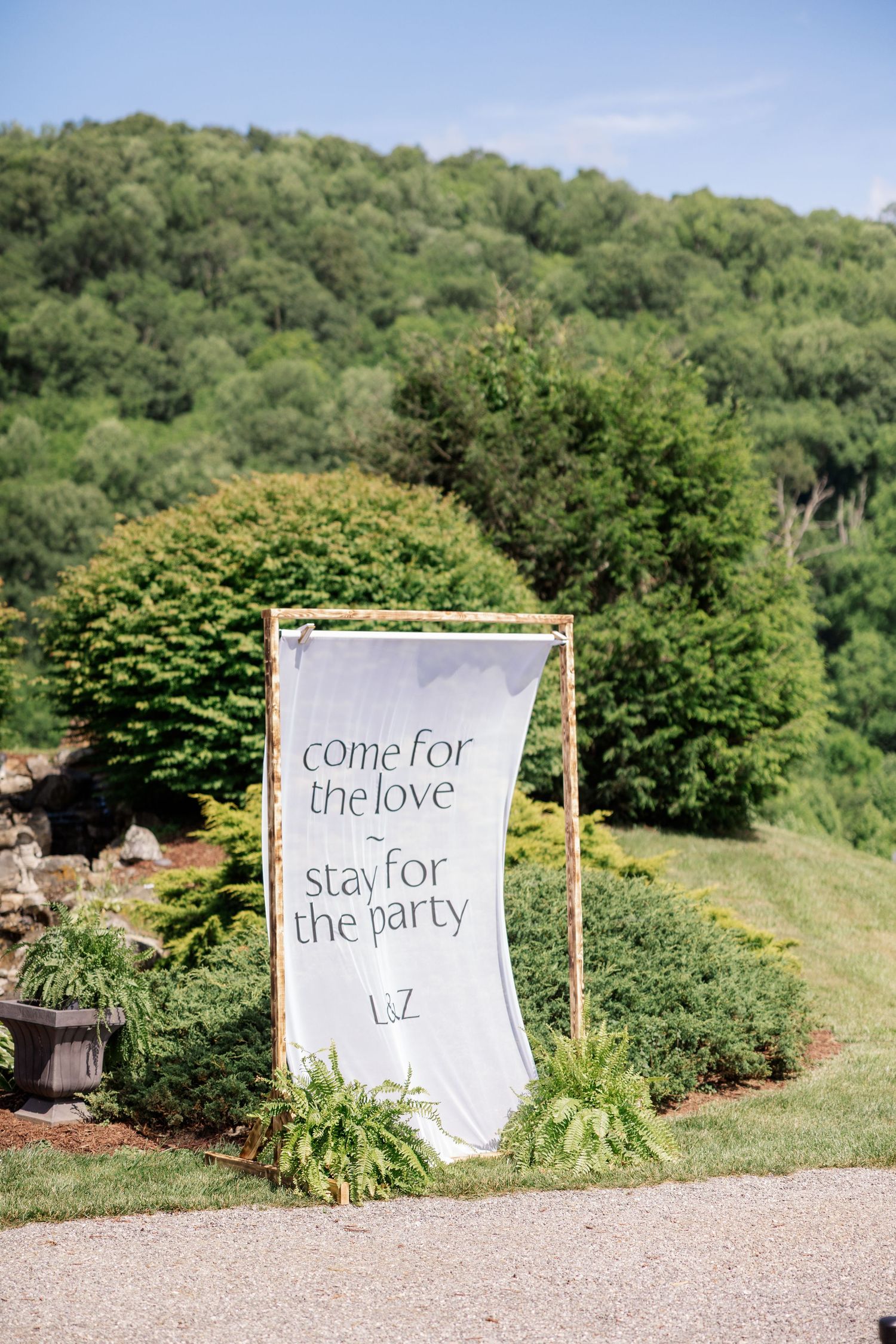 A wedding sign in a lush green landscape reads 'come for the love stay for the party' against mountain backdrop.