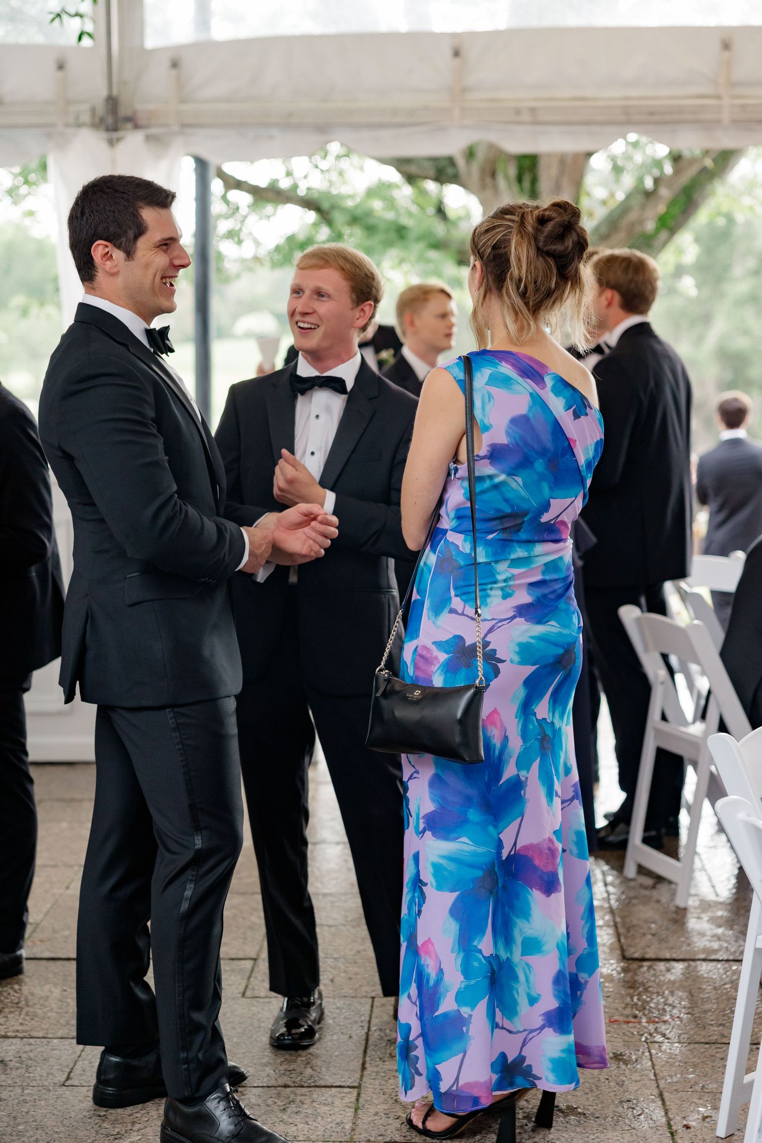 Elegantly dressed wedding guests in formal attire socializing and laughing under a white tent at an outdoor reception.