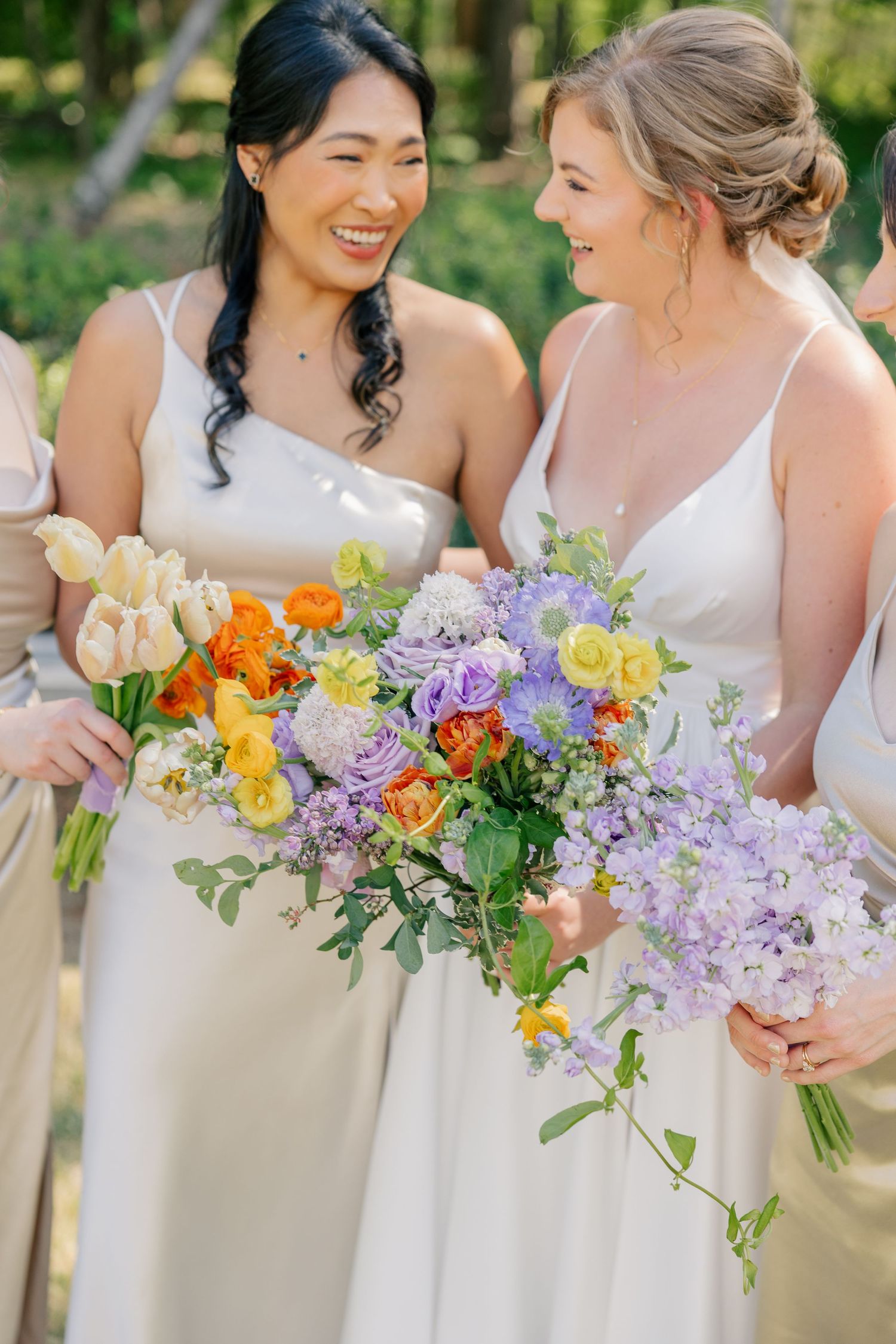 Bridesmaids in champagne gowns laughing and holding colorful spring bouquets with purple, orange, and yellow flowers.