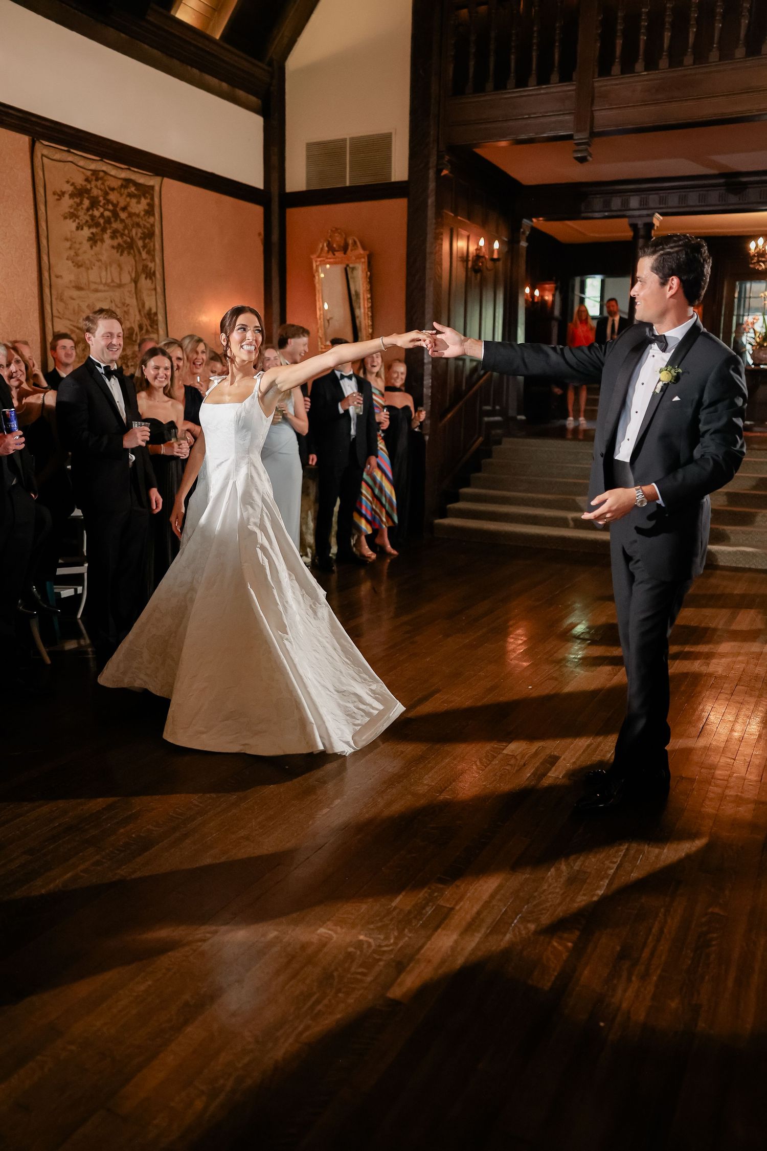 Bride and groom share their first dance in a grand hall while wedding guests look on in formal attire.