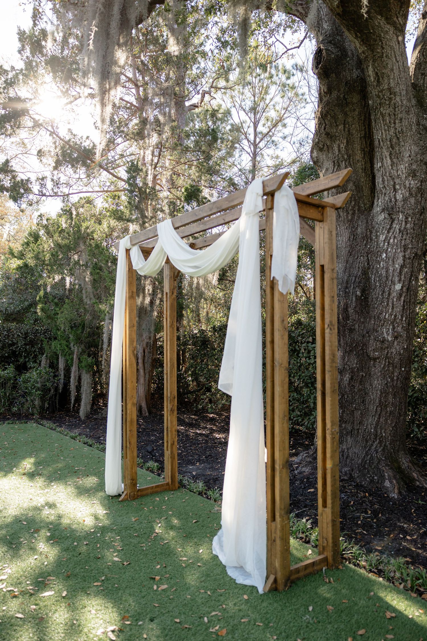 A rustic wooden wedding arch with flowing white fabric draping in a sunlit outdoor garden setting.
