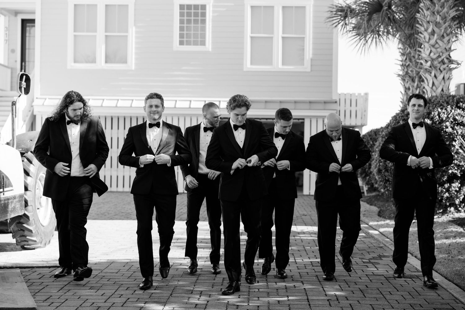 Group of groomsmen in black tuxedos walking and looking down at their phones in a candid moment.