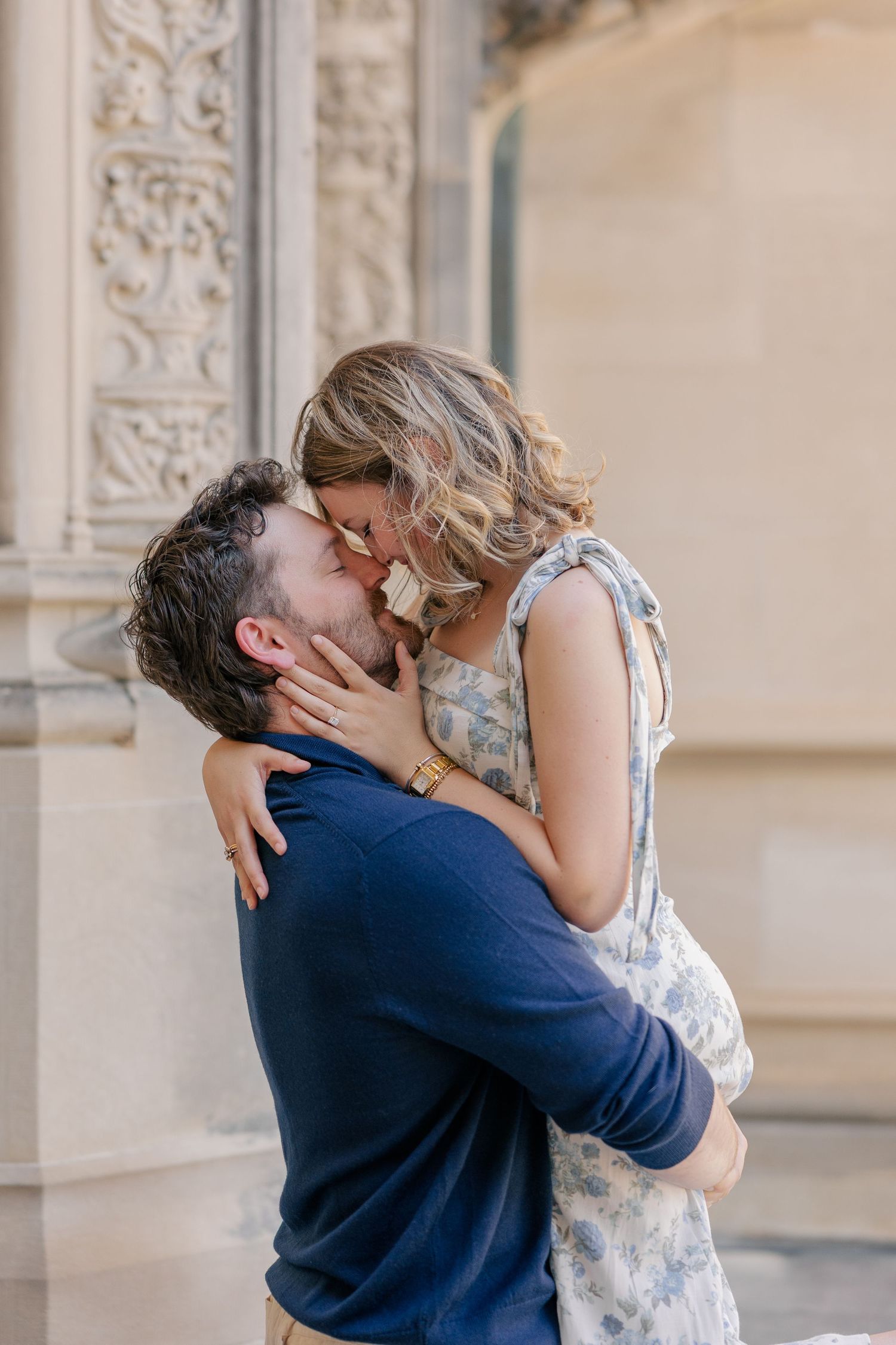 Couple sharing a romantic kiss embrace near ornate stone building exterior in soft natural light.