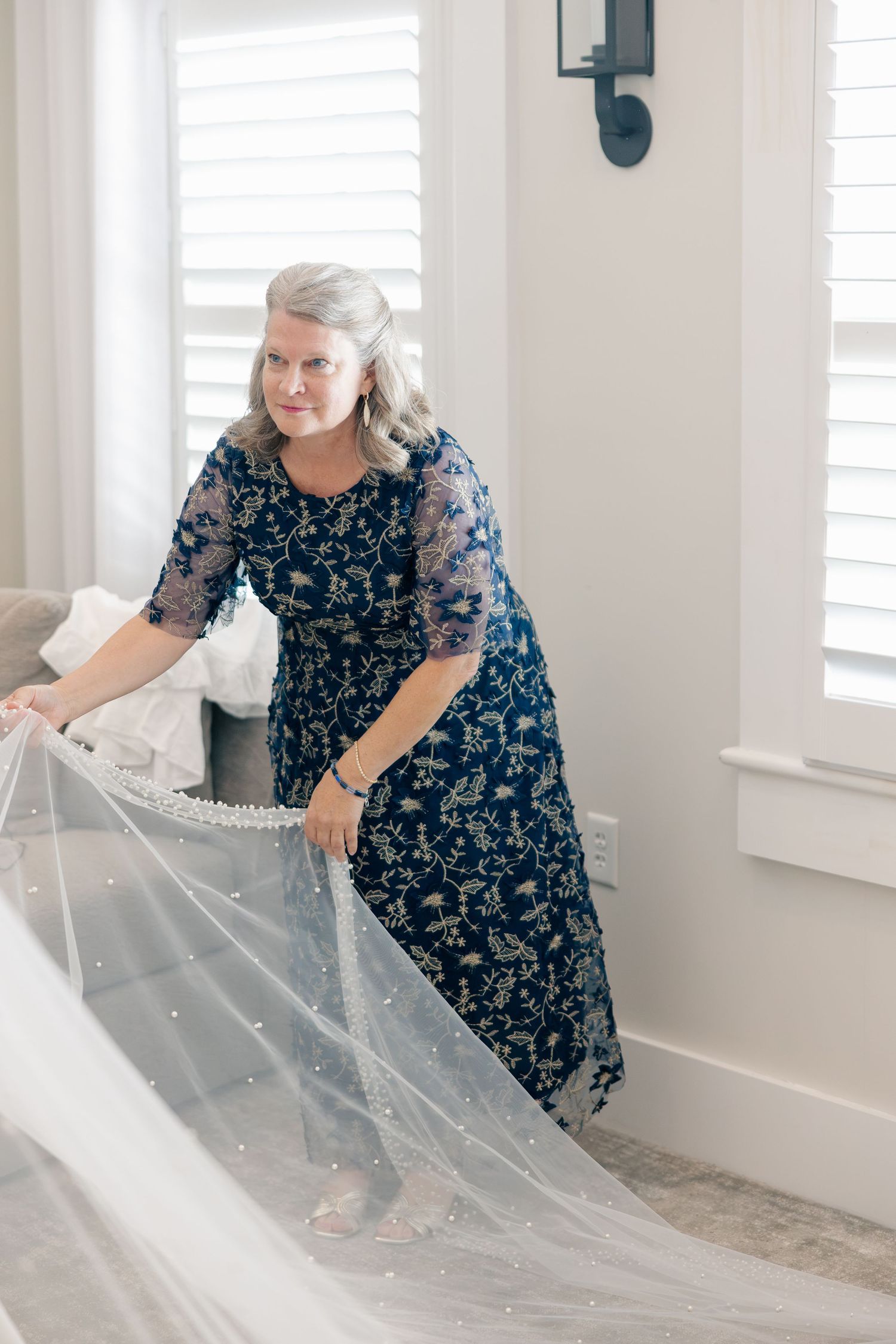 Elegant mother of the bride in navy floral gown arranging a pearl-embellished wedding veil in a bright white room.