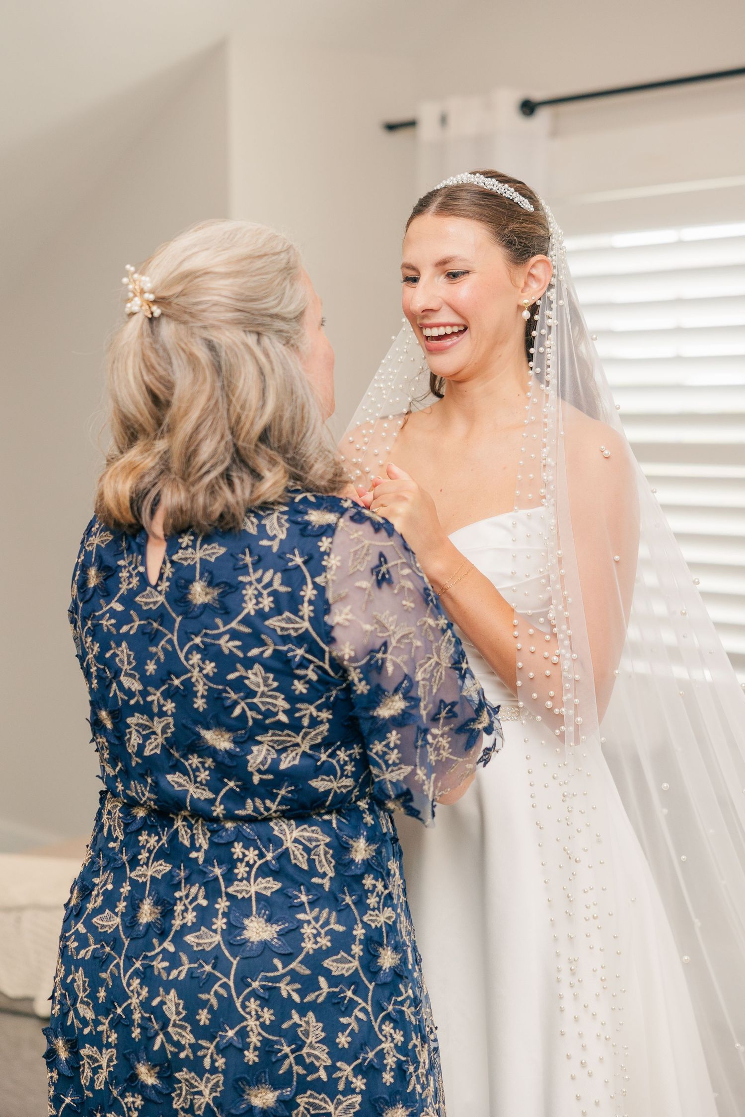 Bride in white gown and veil laughing with older woman in blue floral dress during wedding preparation.