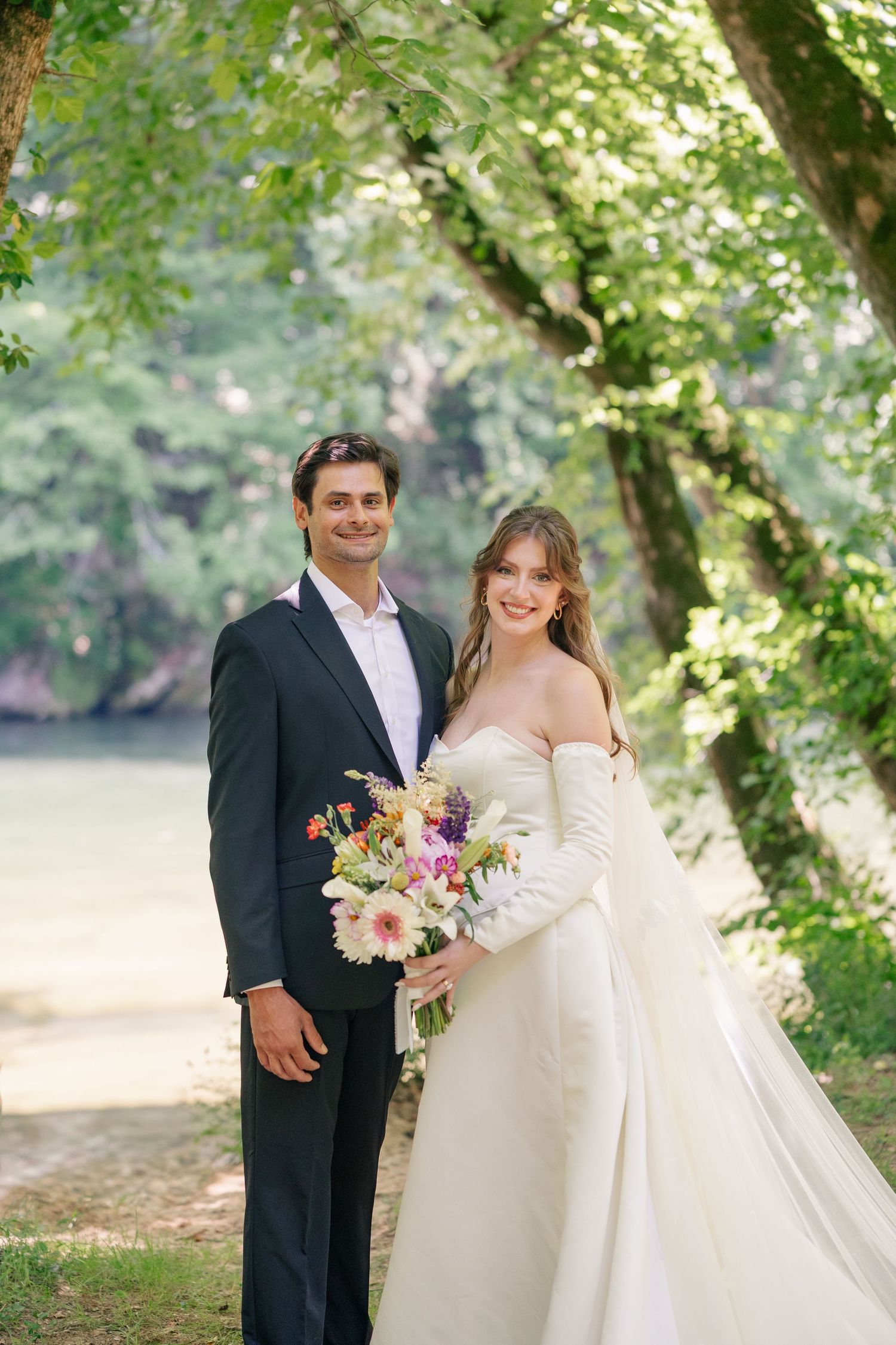 Bride and groom smiling together outdoors near a river, she holds a colorful bouquet in a white off-shoulder gown.